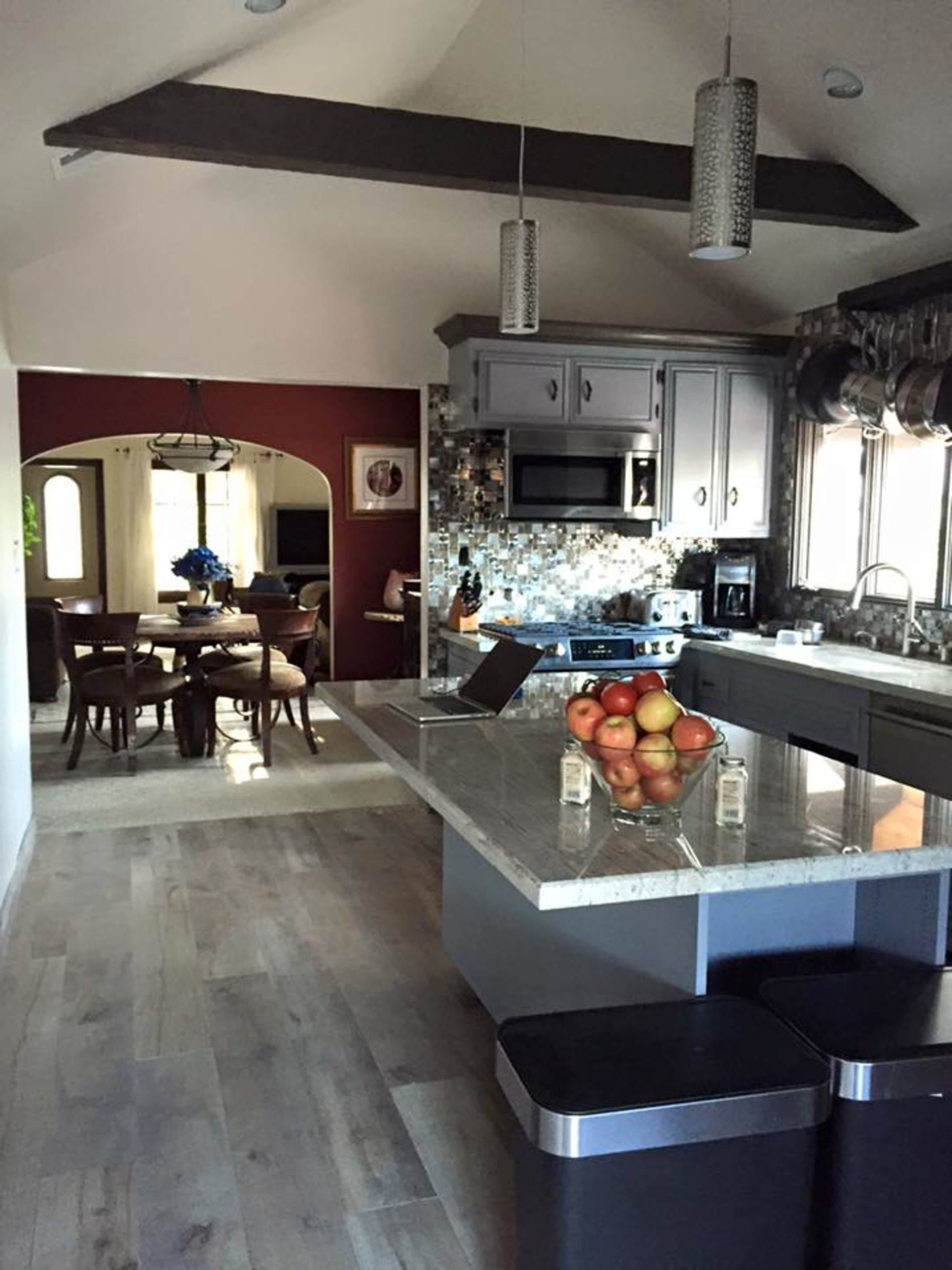 A modern kitchen features a large island with a bowl of apples, leading into a dining area with a round table and chairs.