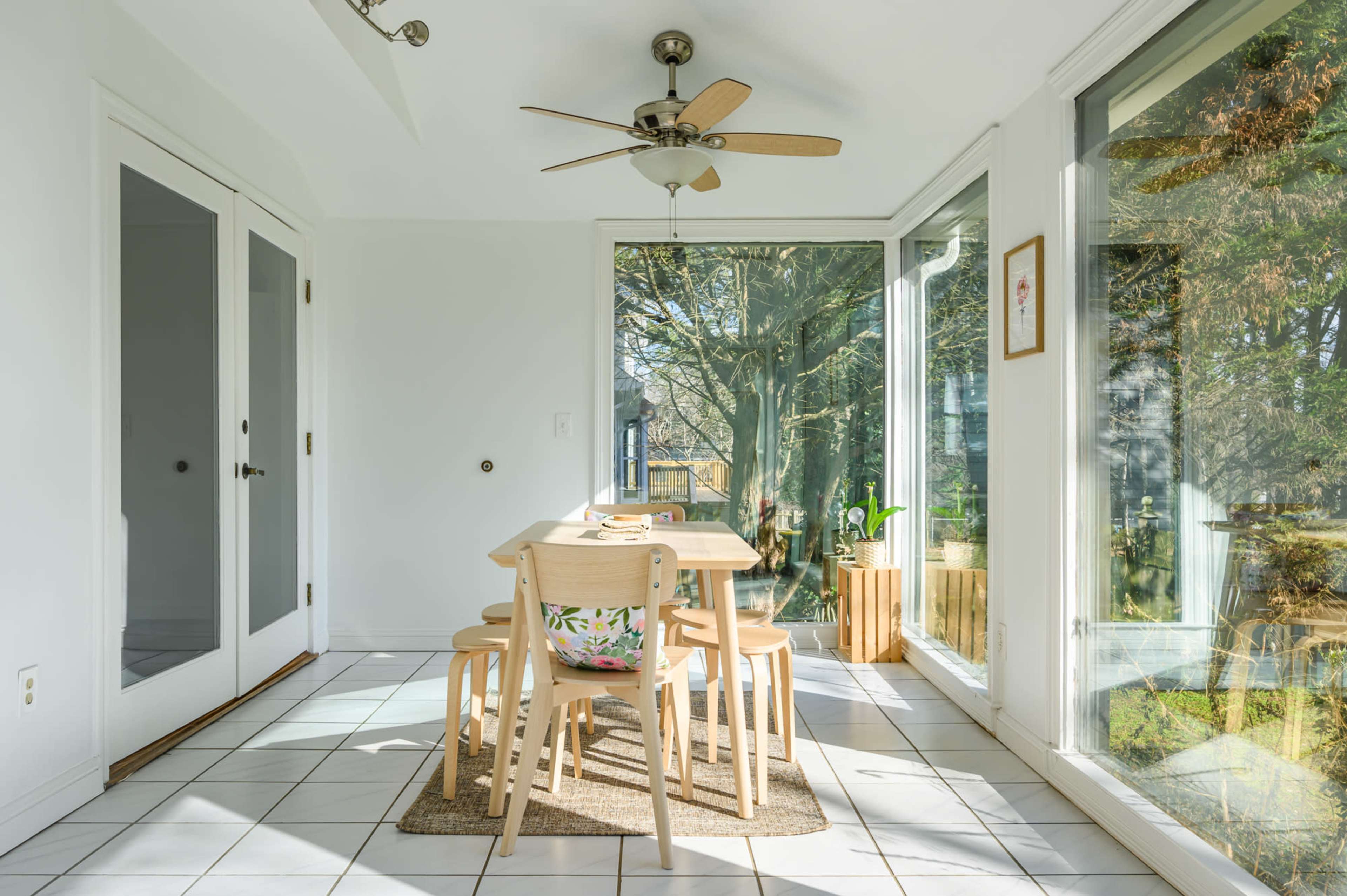 A bright dining area features a table with four chairs near large windows surrounded by greenery.