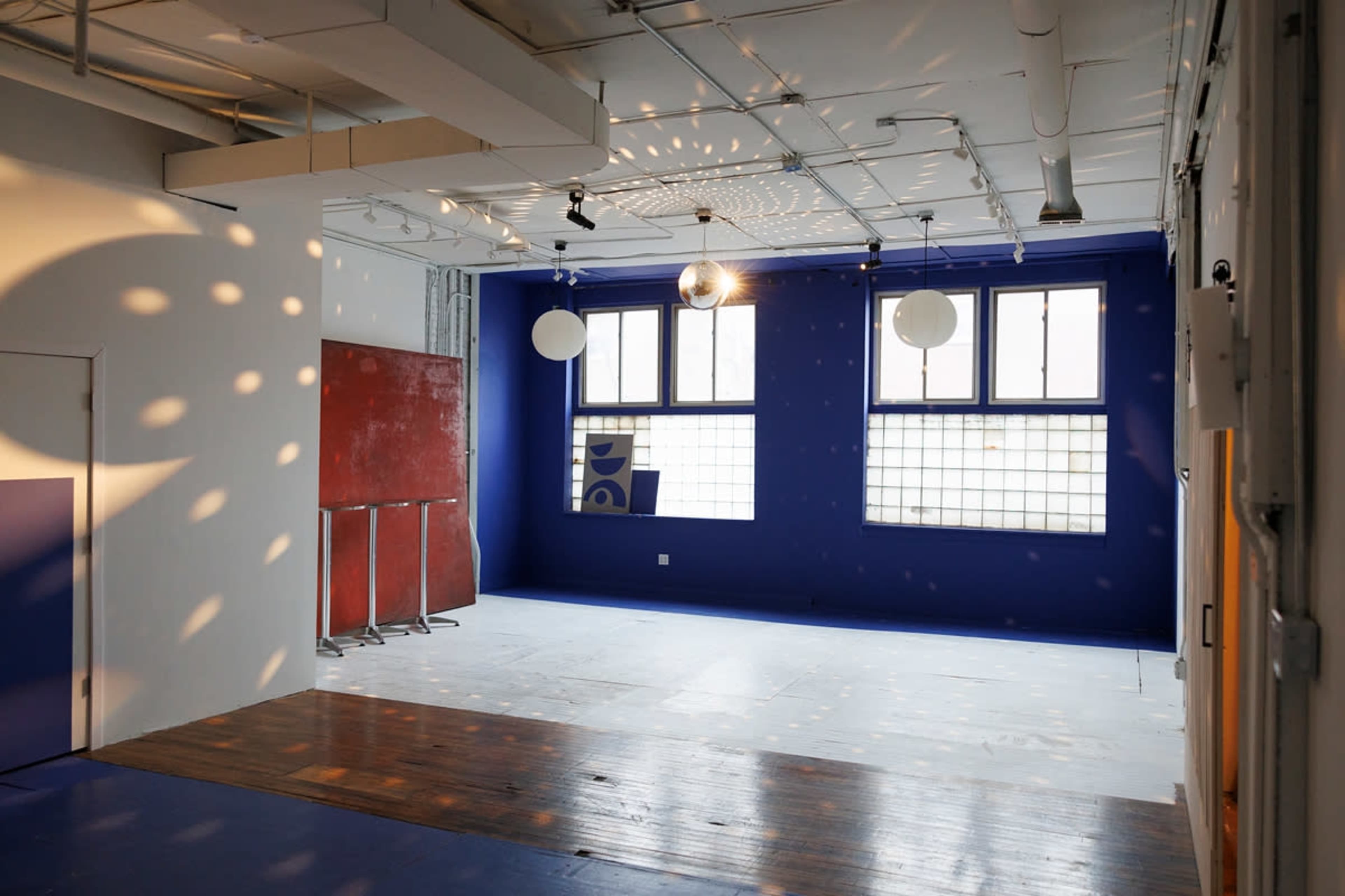 The image shows a spacious, brightly lit room with blue walls, wooden flooring, and two large windows.