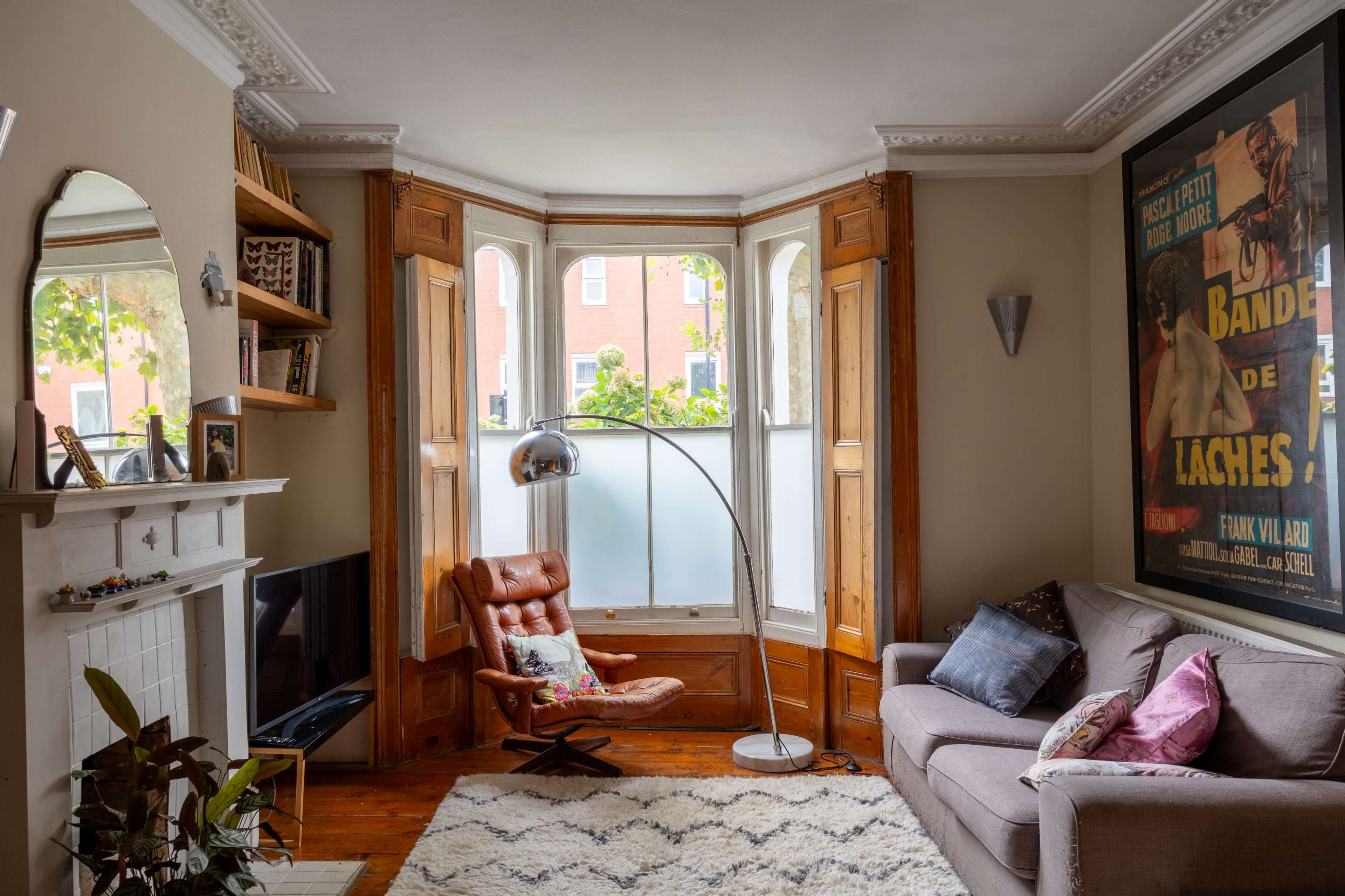 A cozy living room features a brown leather armchair, a gray sofa, a large window with a view of greenery, and a prominent movie poster on the wall.