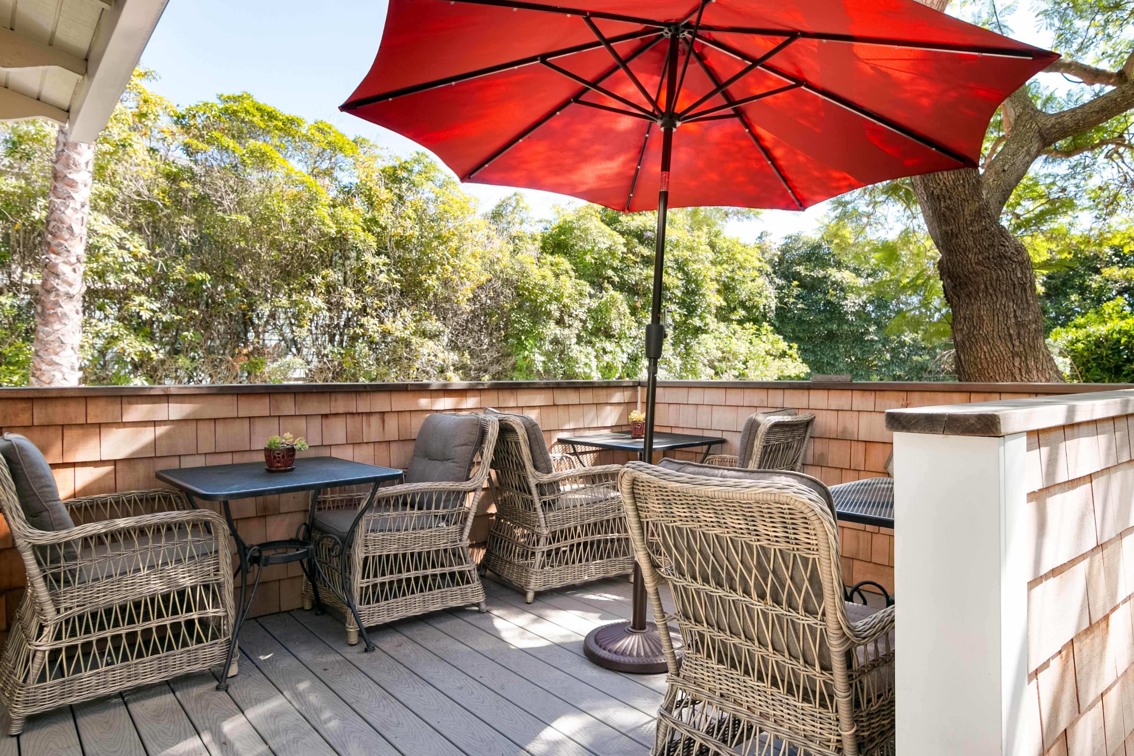 A patio with a red umbrella shades wicker furniture arranged around small tables, surrounded by greenery.
