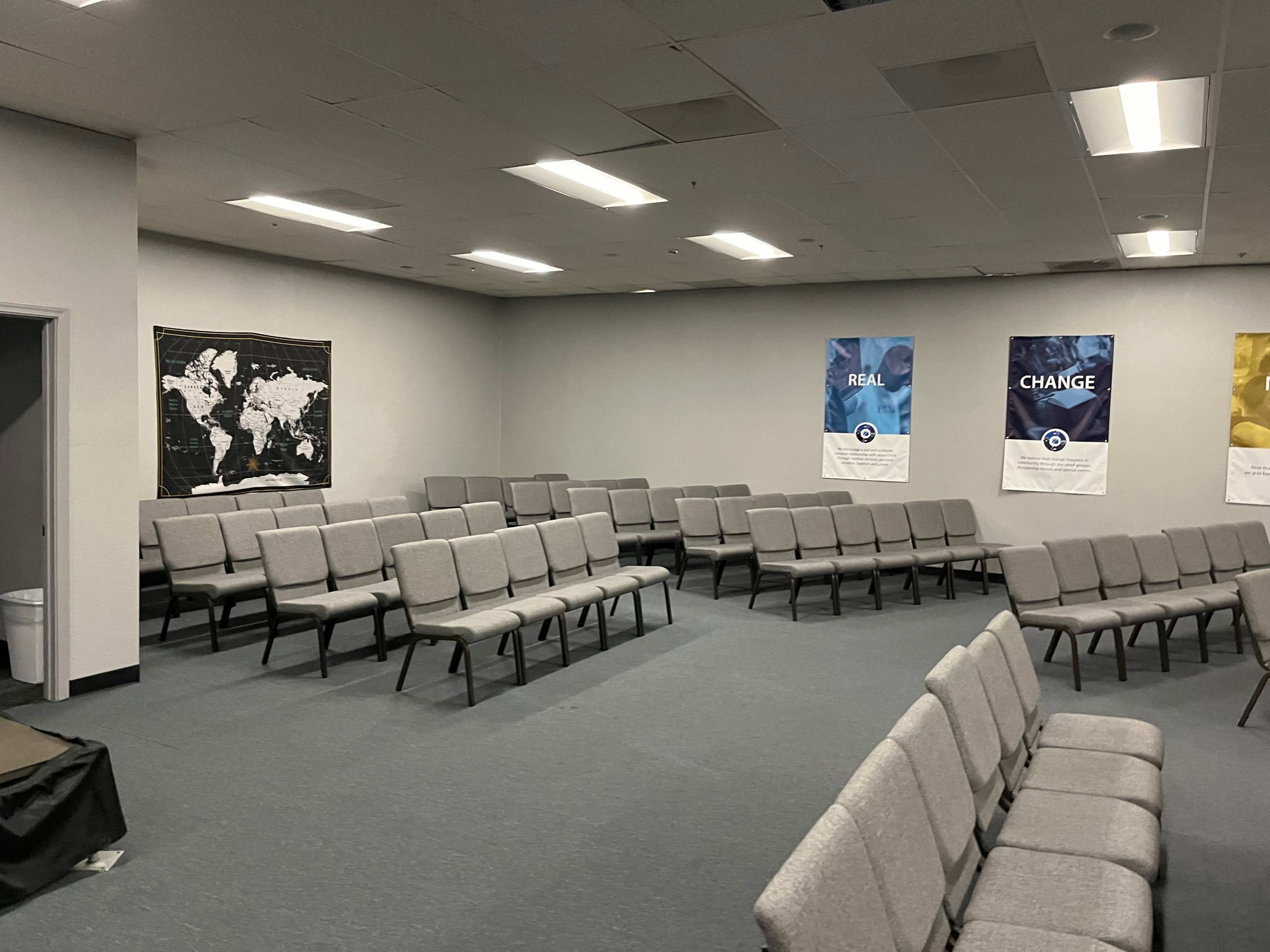 A sparsely furnished room with rows of gray chairs arranged for a meeting, a world map on one wall, and informational banners hanging on another.