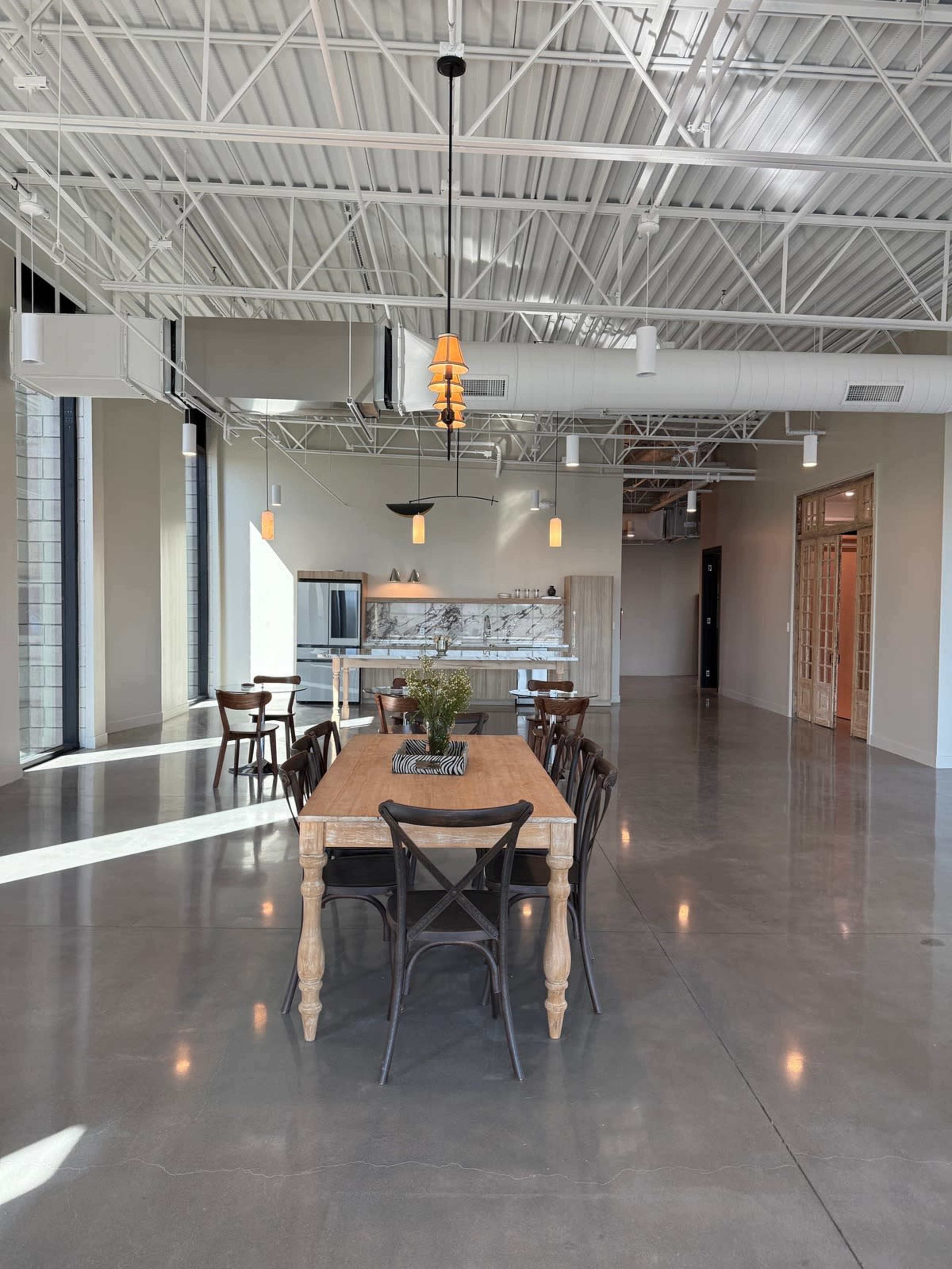 The image shows a spacious, modern dining area with a wooden table surrounded by chairs, adjacent to a kitchen space with light-colored cabinetry and a counter.