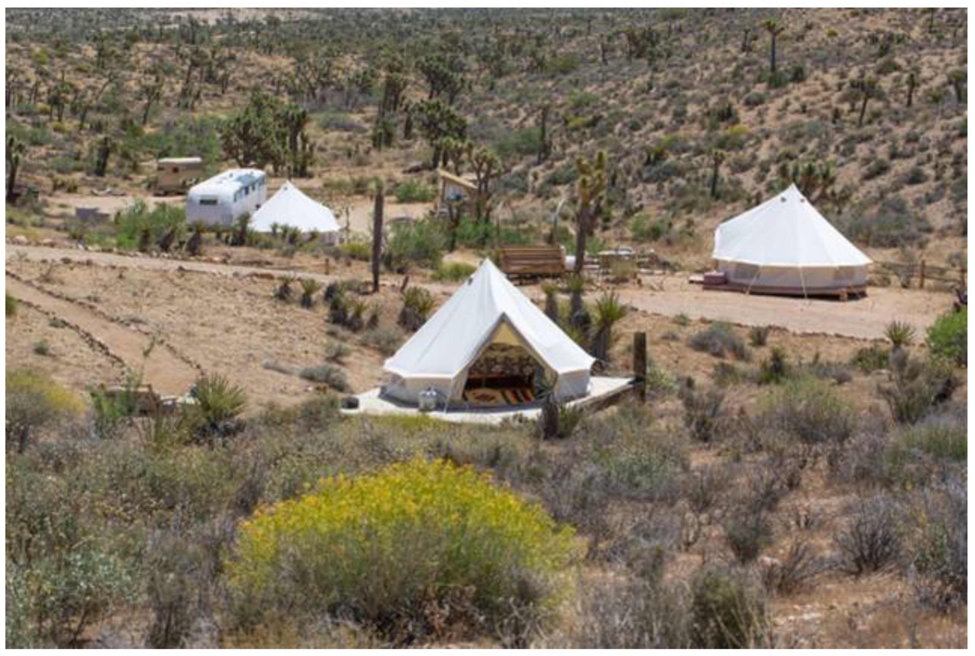 The image shows several white tents and a trailer set up in a desert landscape with sparse vegetation and rolling hills in the background.
