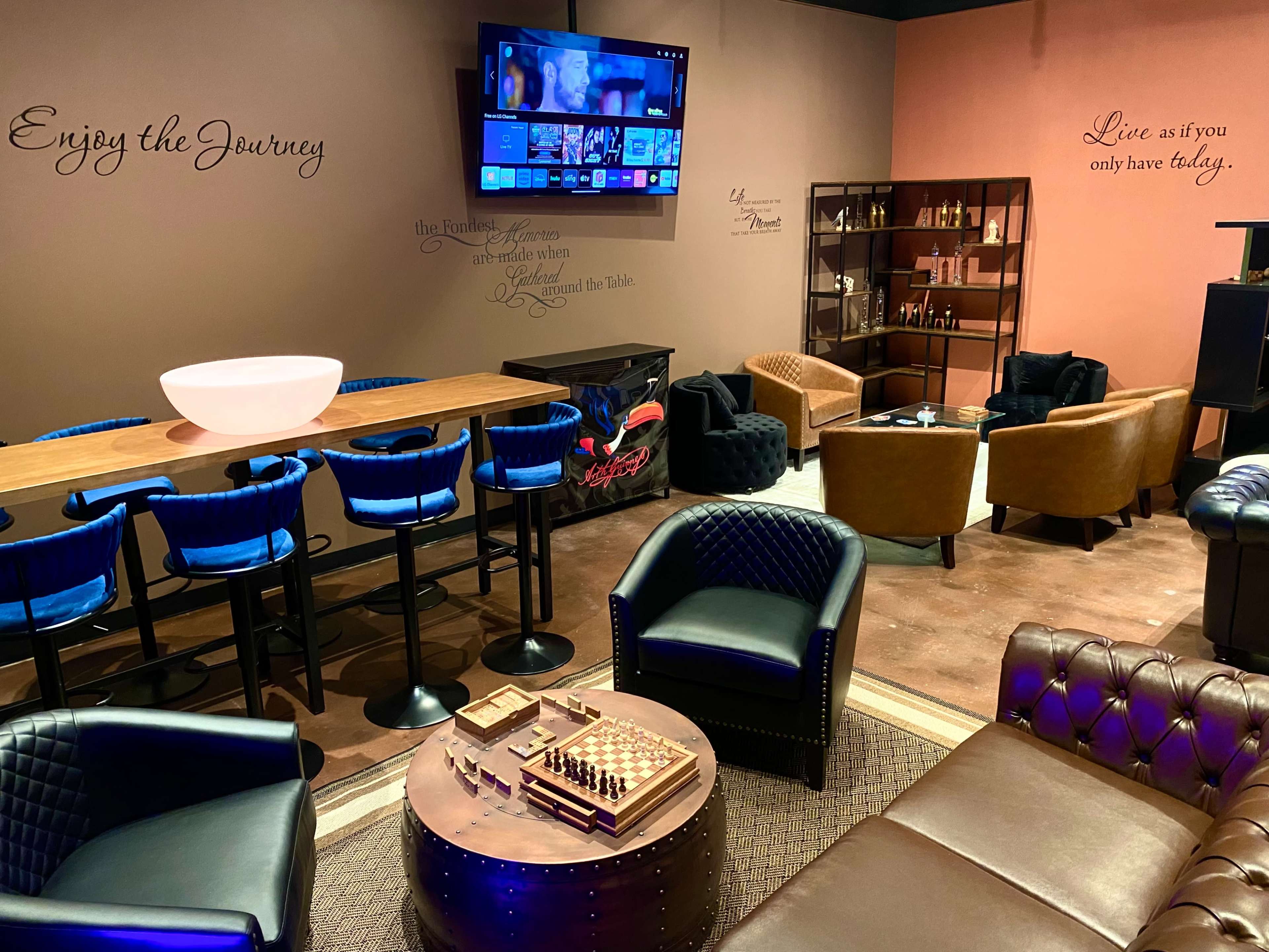 The image shows a stylish lounge area featuring a bar with stools, a large television, comfortable seating, and decorative wall inscriptions.