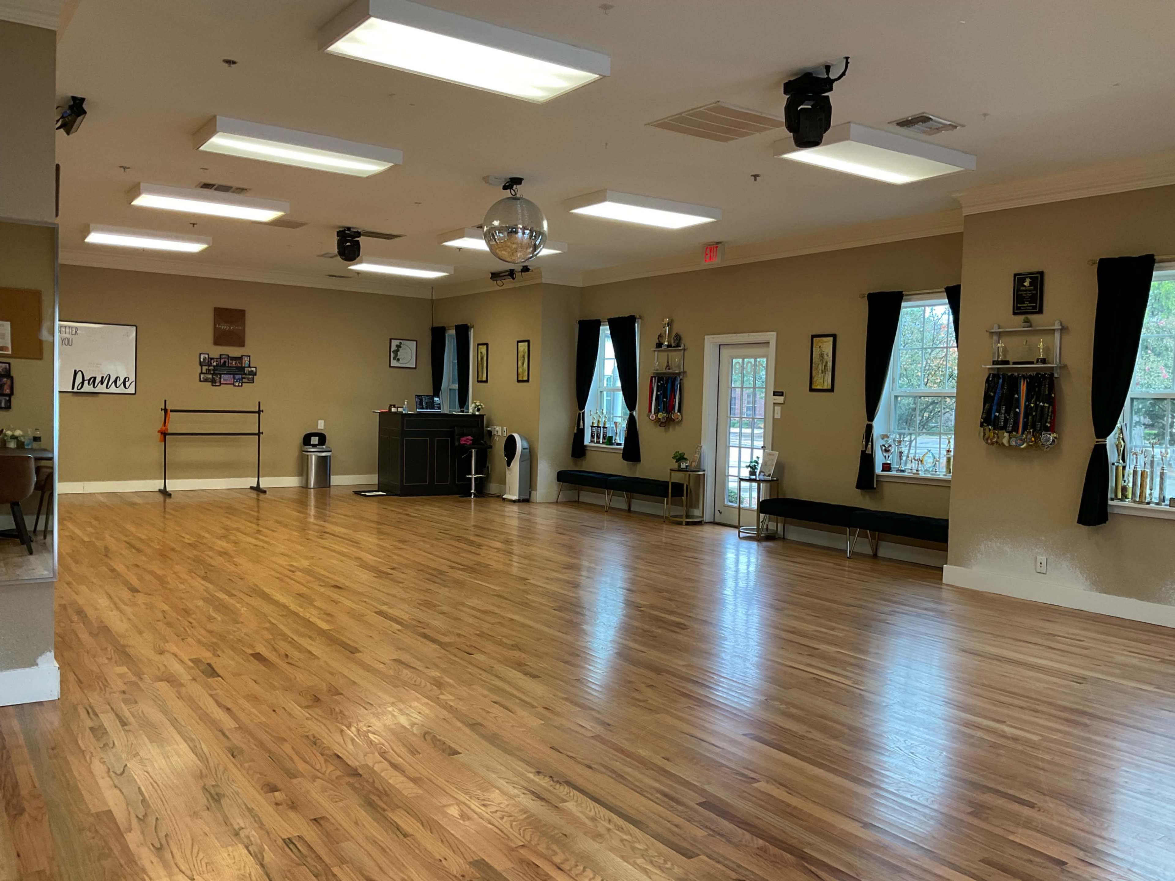 The image shows a spacious dance studio with wooden flooring, mirrors on the walls, and a reception area in the background.