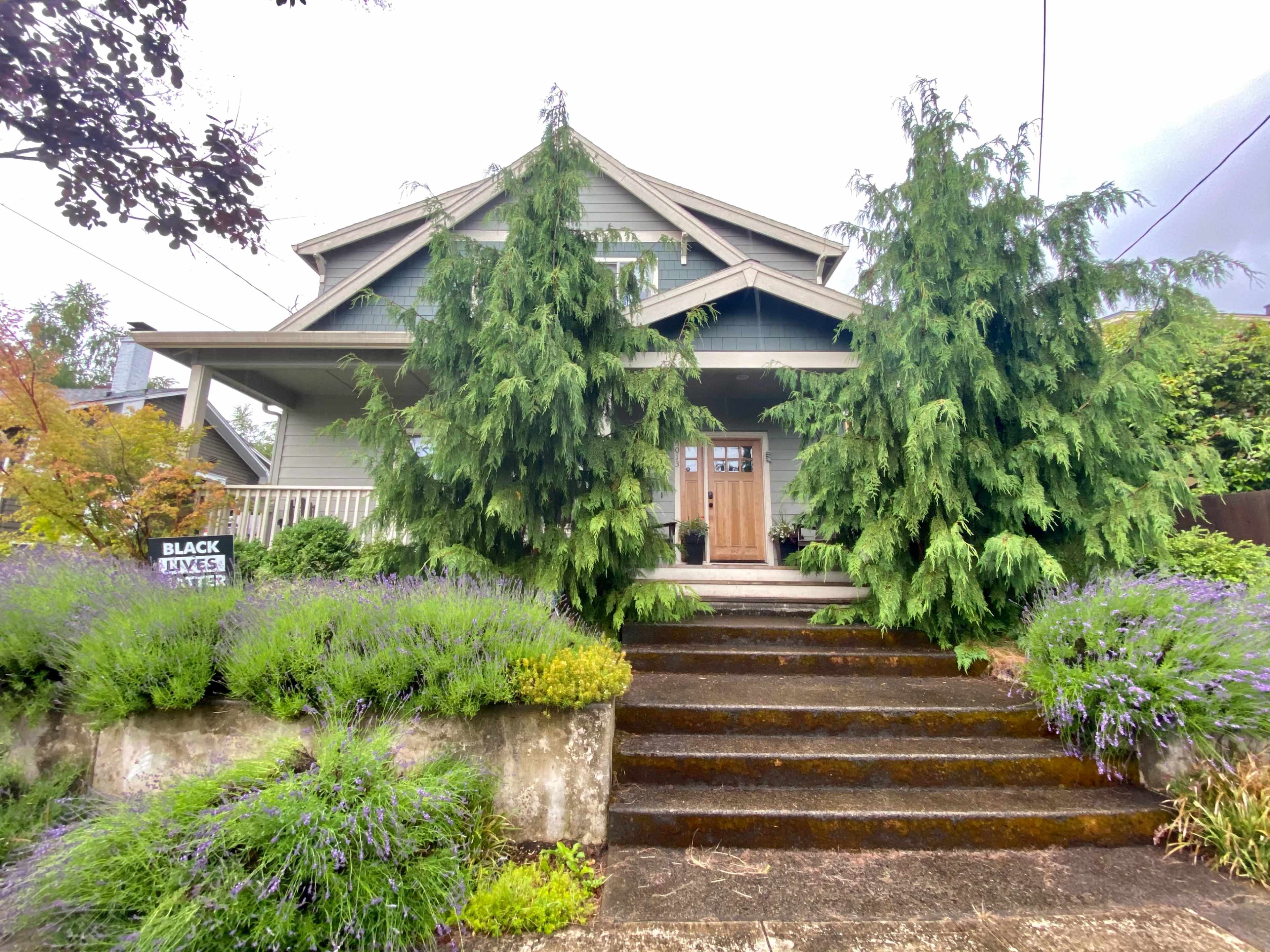 A two-story green house is flanked by tall, bushy trees and surrounded by colorful landscaping, with a visible sign that reads "BLACK LIVES MATTER."