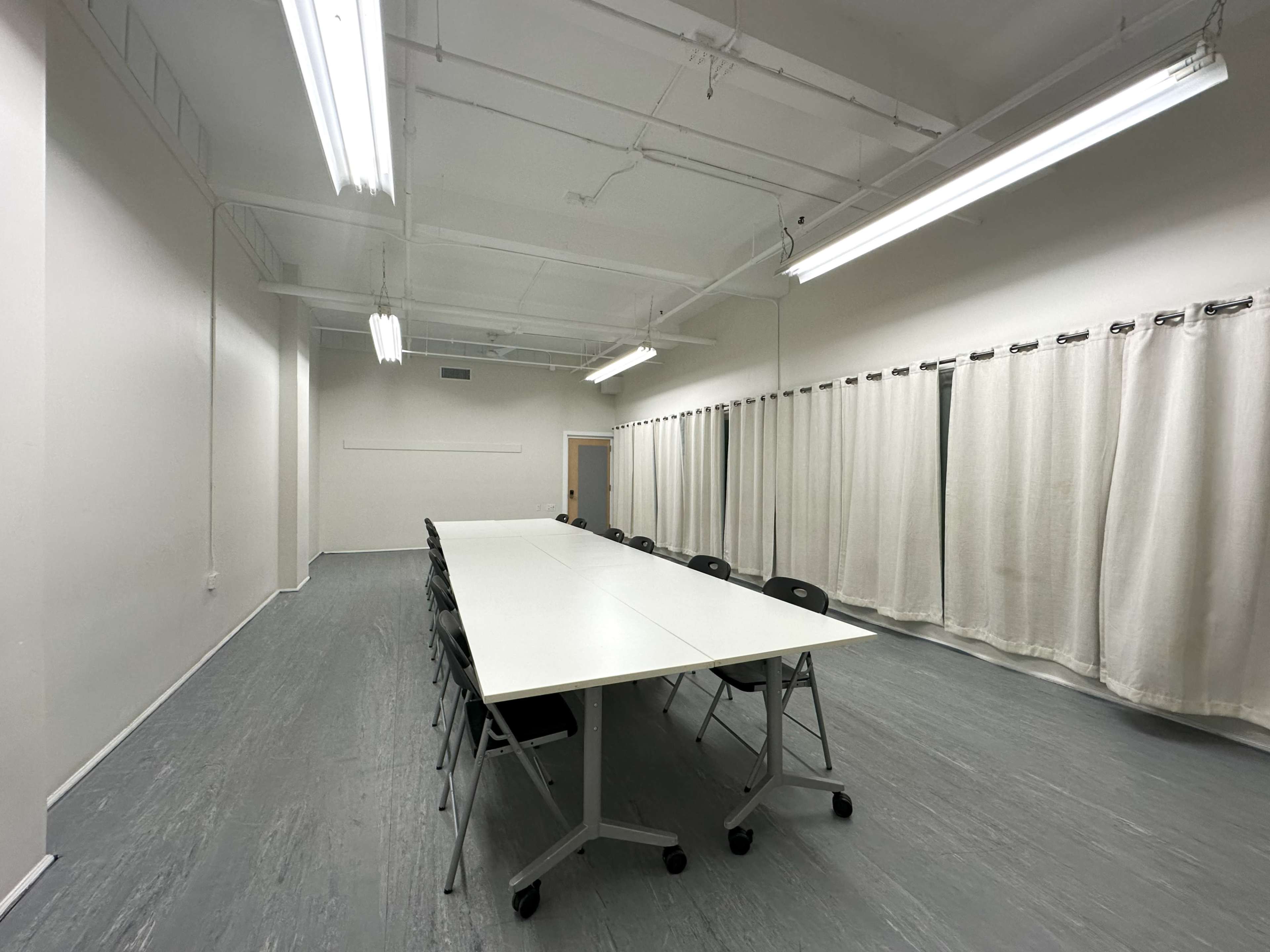 A long, white table is surrounded by black chairs in a brightly lit, empty meeting room with beige curtains.