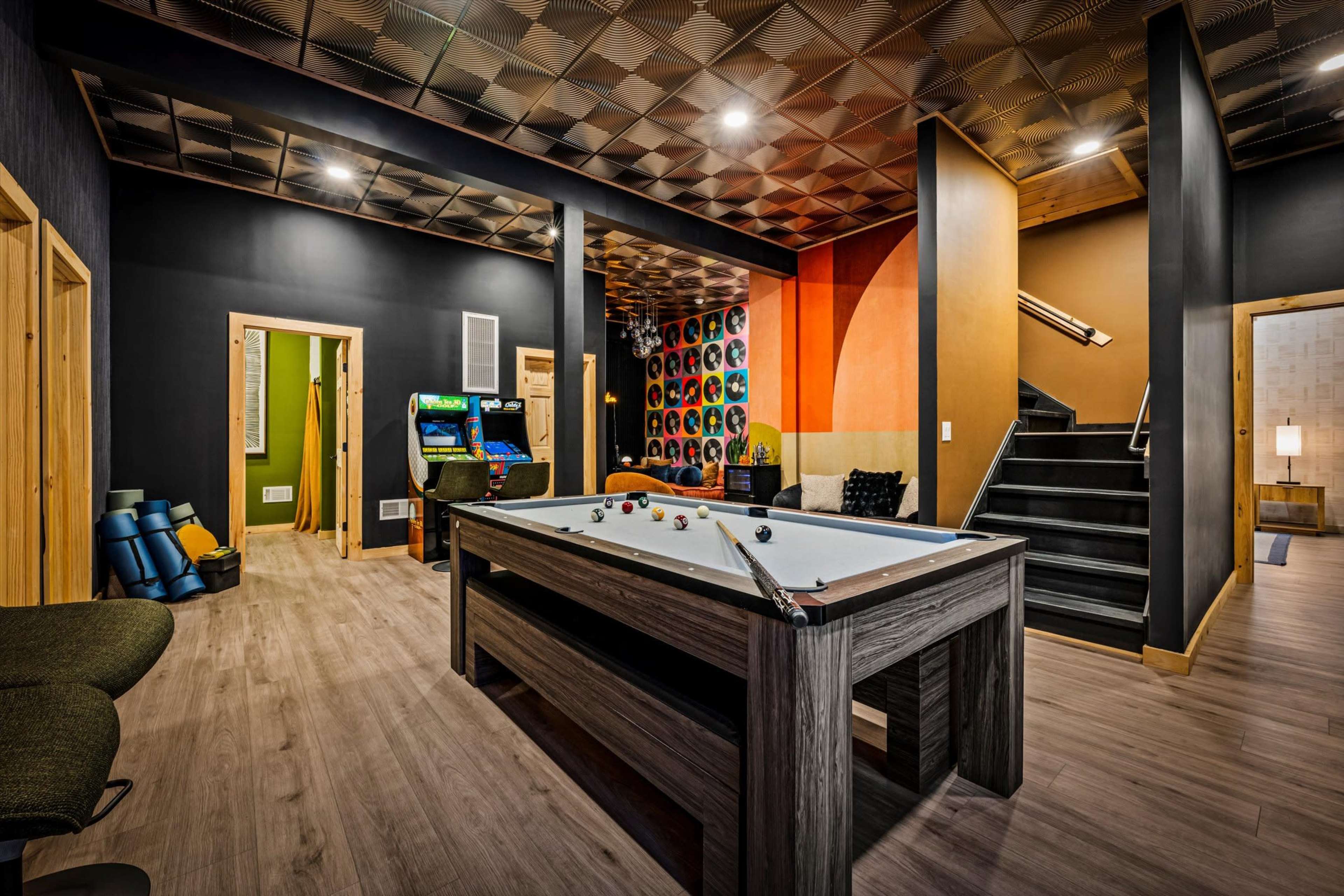 The image shows a recreational room featuring a billiards table, arcade game, and access to a staircase, with vibrant wall colors and modern decor.