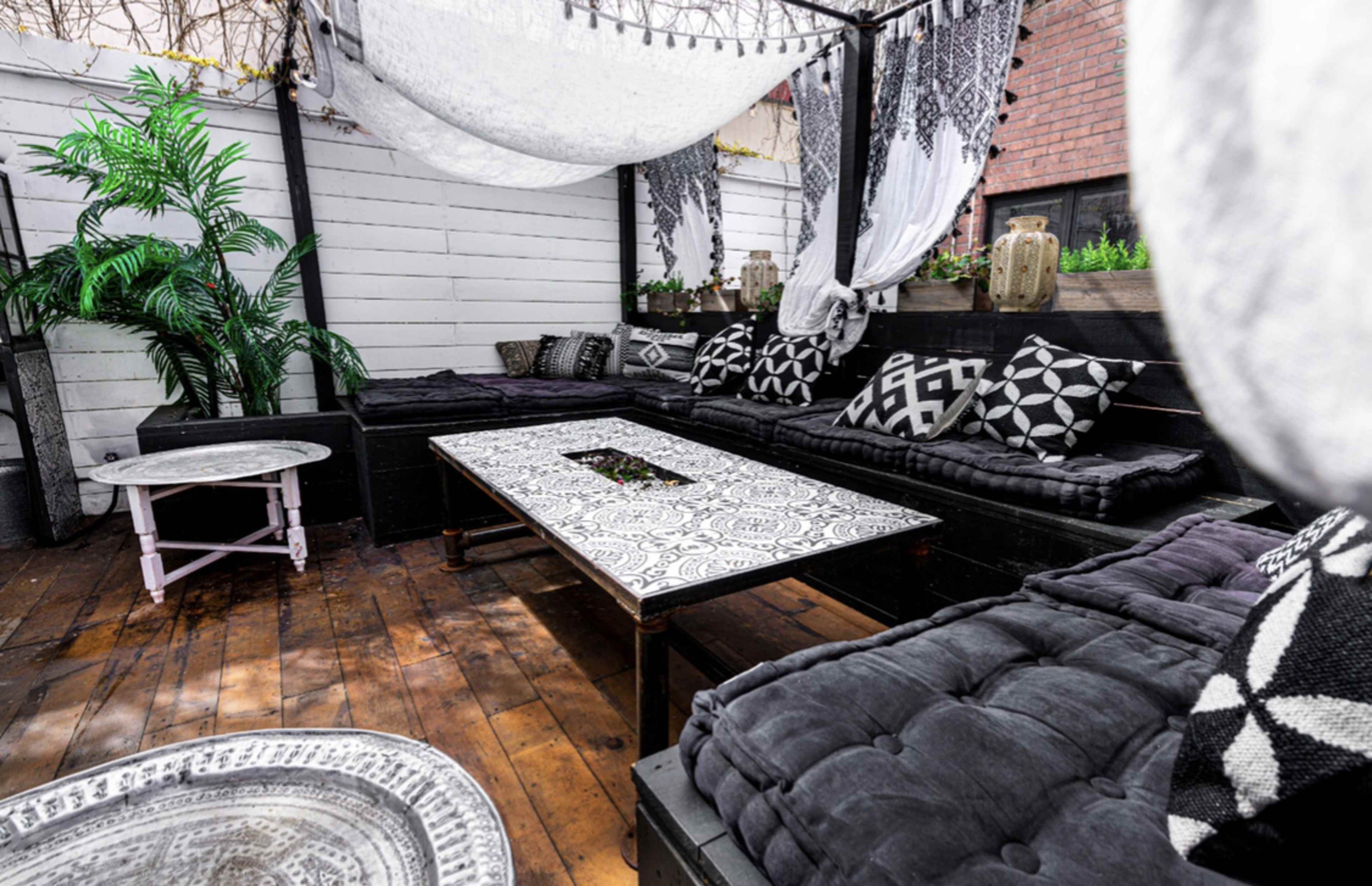 The image shows a cozy outdoor seating area featuring black cushions, patterned throw pillows, and a decorated table under a draped canopy.