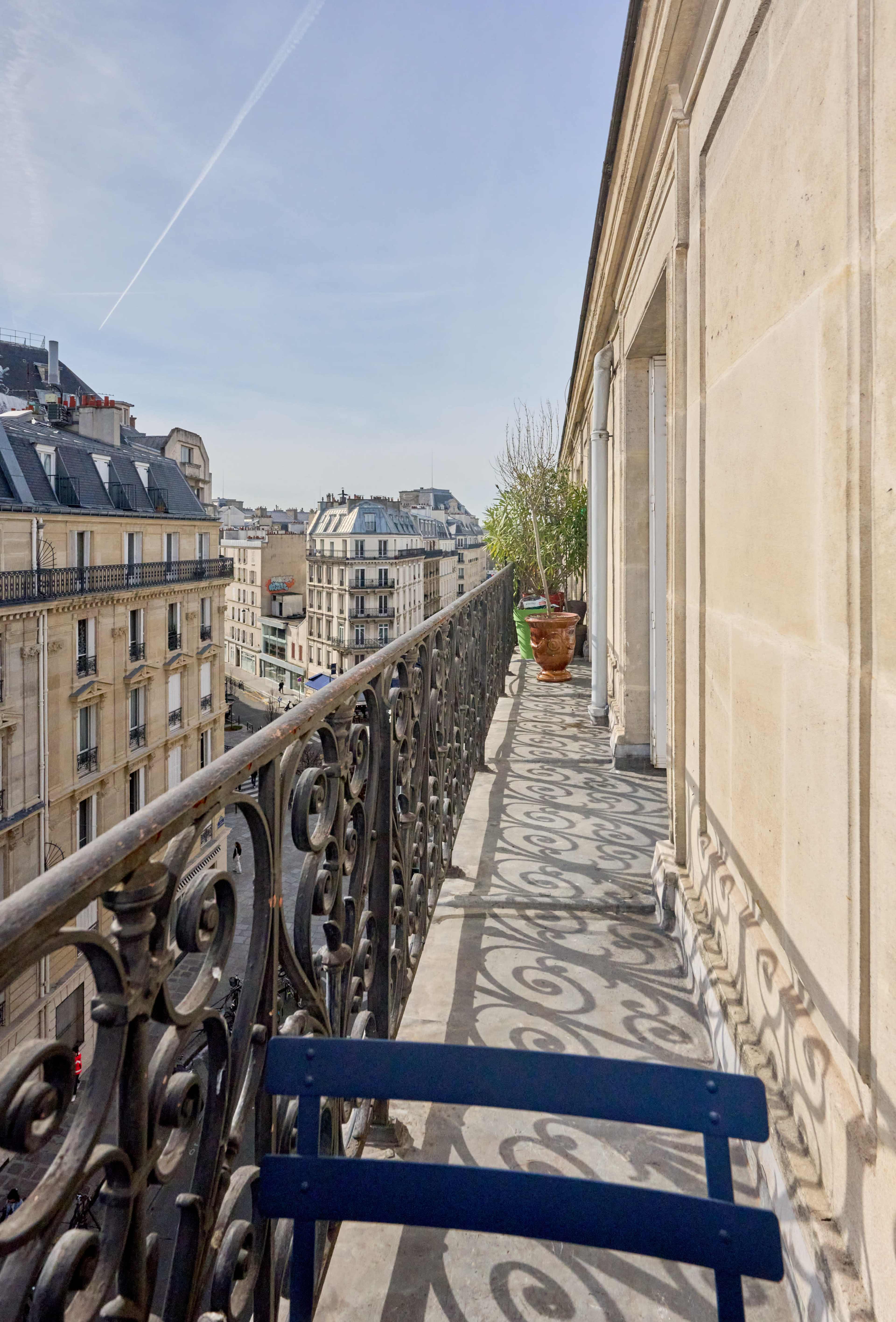 A balcony with a wrought-iron railing overlooks a cityscape that includes buildings and a clear sky.