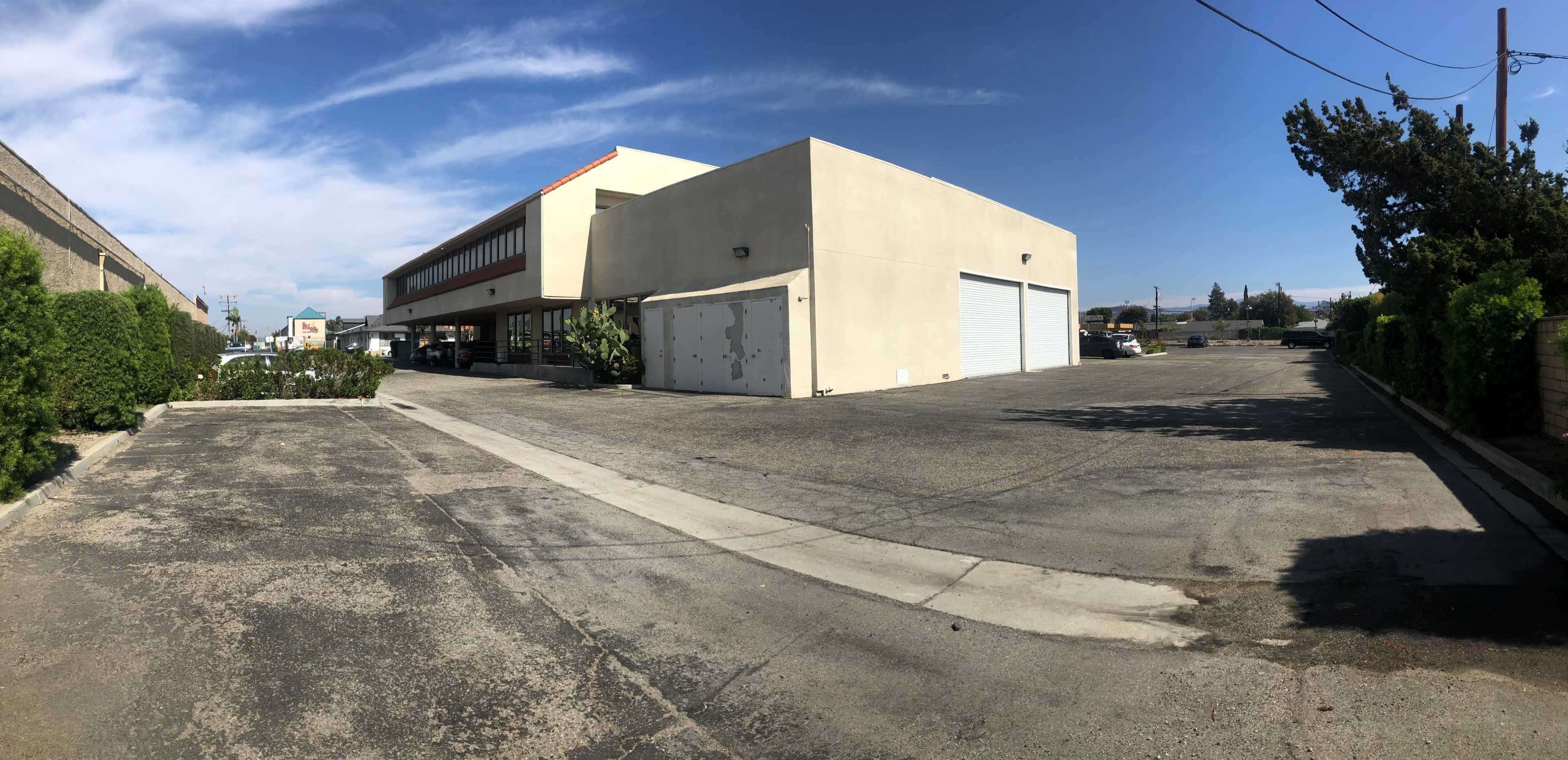 The image shows a large, beige commercial building with a garage door and a paved parking area next to it, under a blue sky with some clouds.