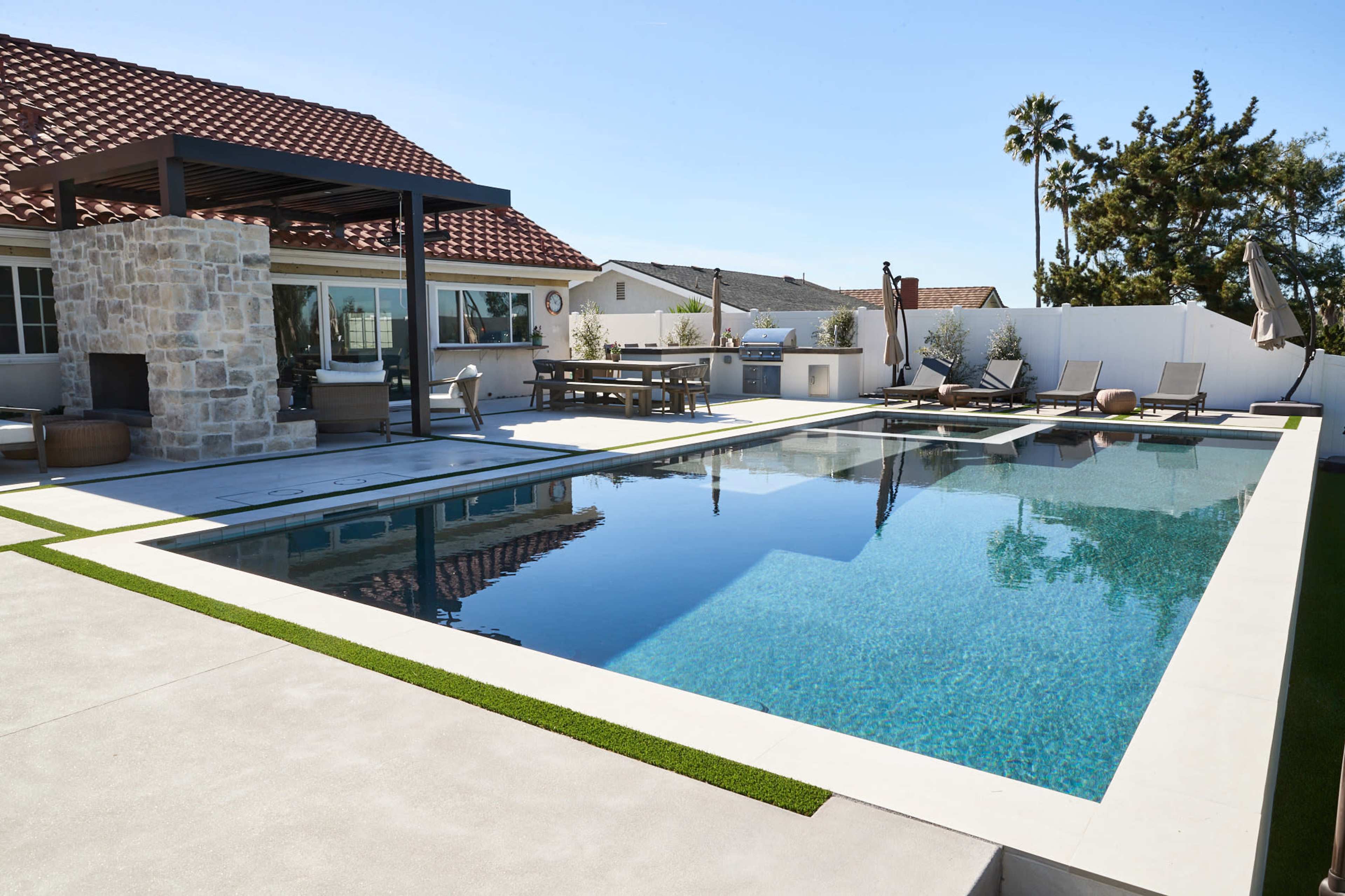 A modern outdoor pool area with a stone fireplace, lounge chairs, and a shaded dining space under a pergola.