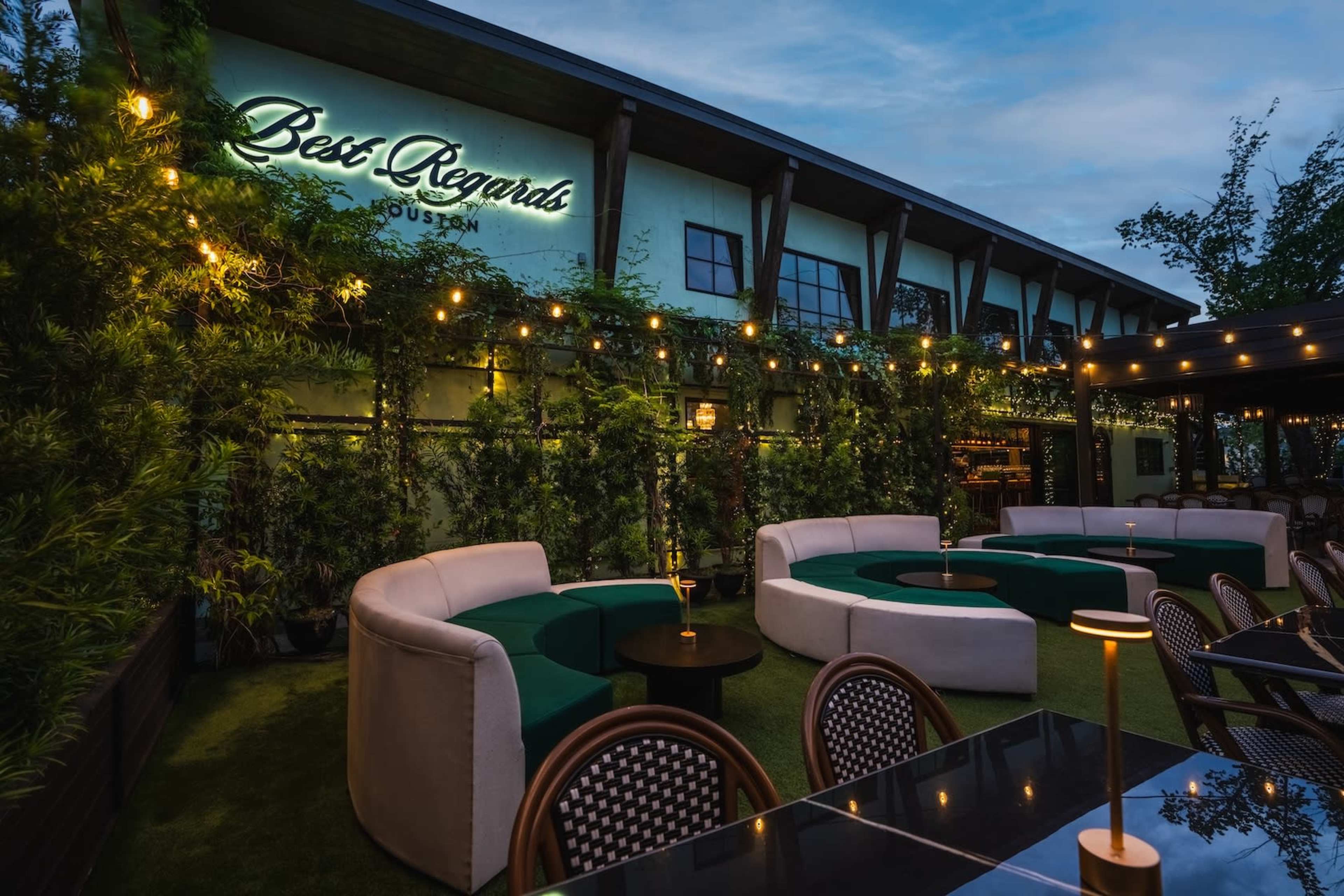 A patio area features circular seating arrangements with green cushions and soft lighting, adjacent to a building displaying a sign that reads "Best Regards."