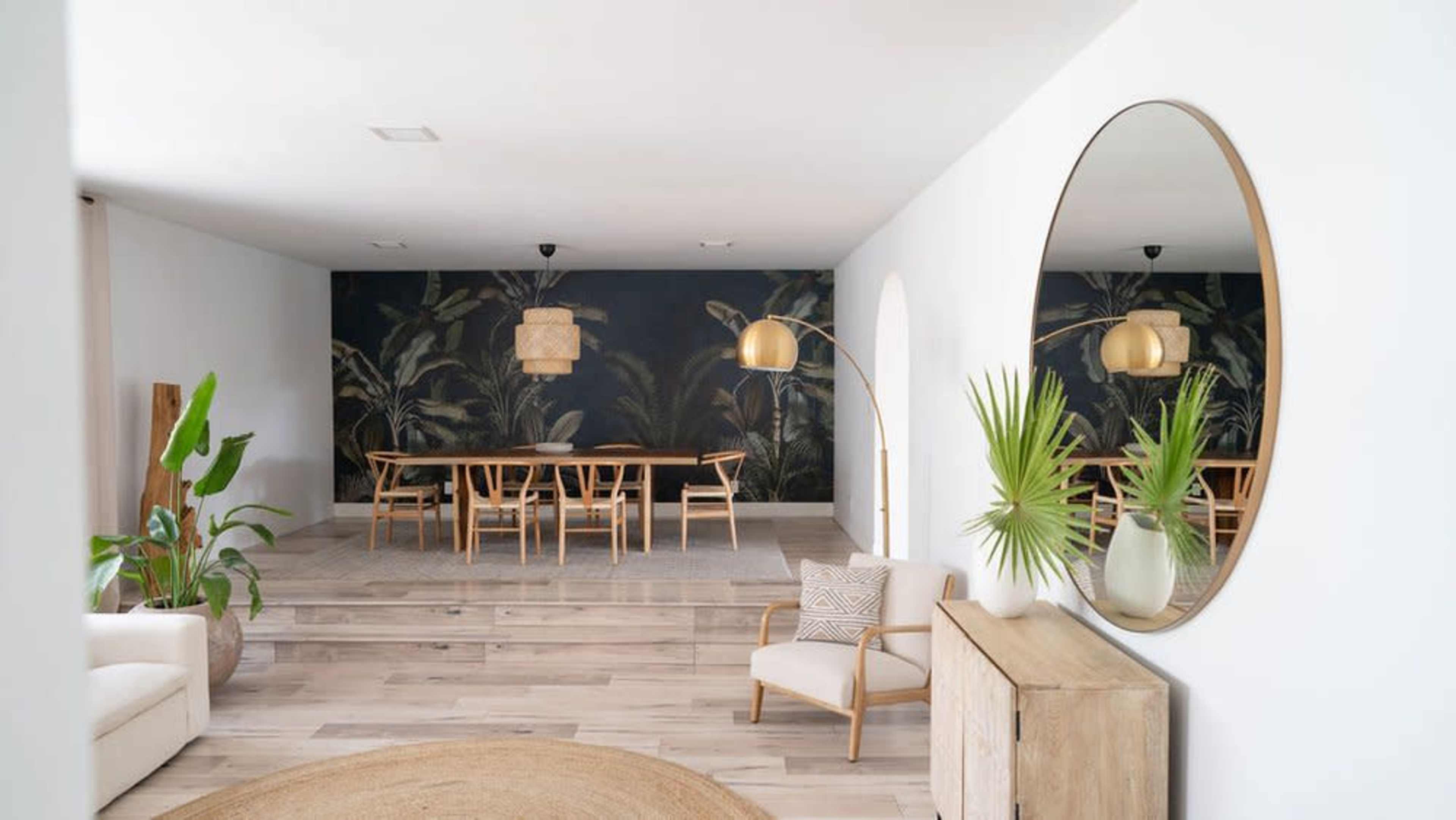 The image shows a bright, modern interior featuring a dining area with a wooden table, chairs, and a wall decorated with a tropical mural, along with wooden flooring and a round mirror on the wall.