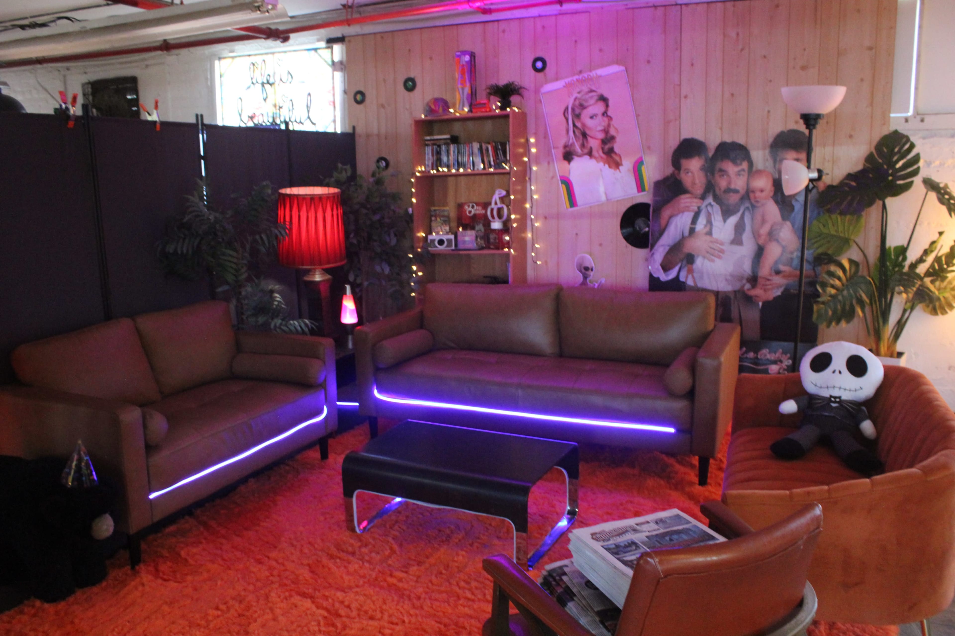 The image shows a cozy lounge area with two brown sofas, a coffee table, and colorful decorative elements, including a red lamp and various plants.
