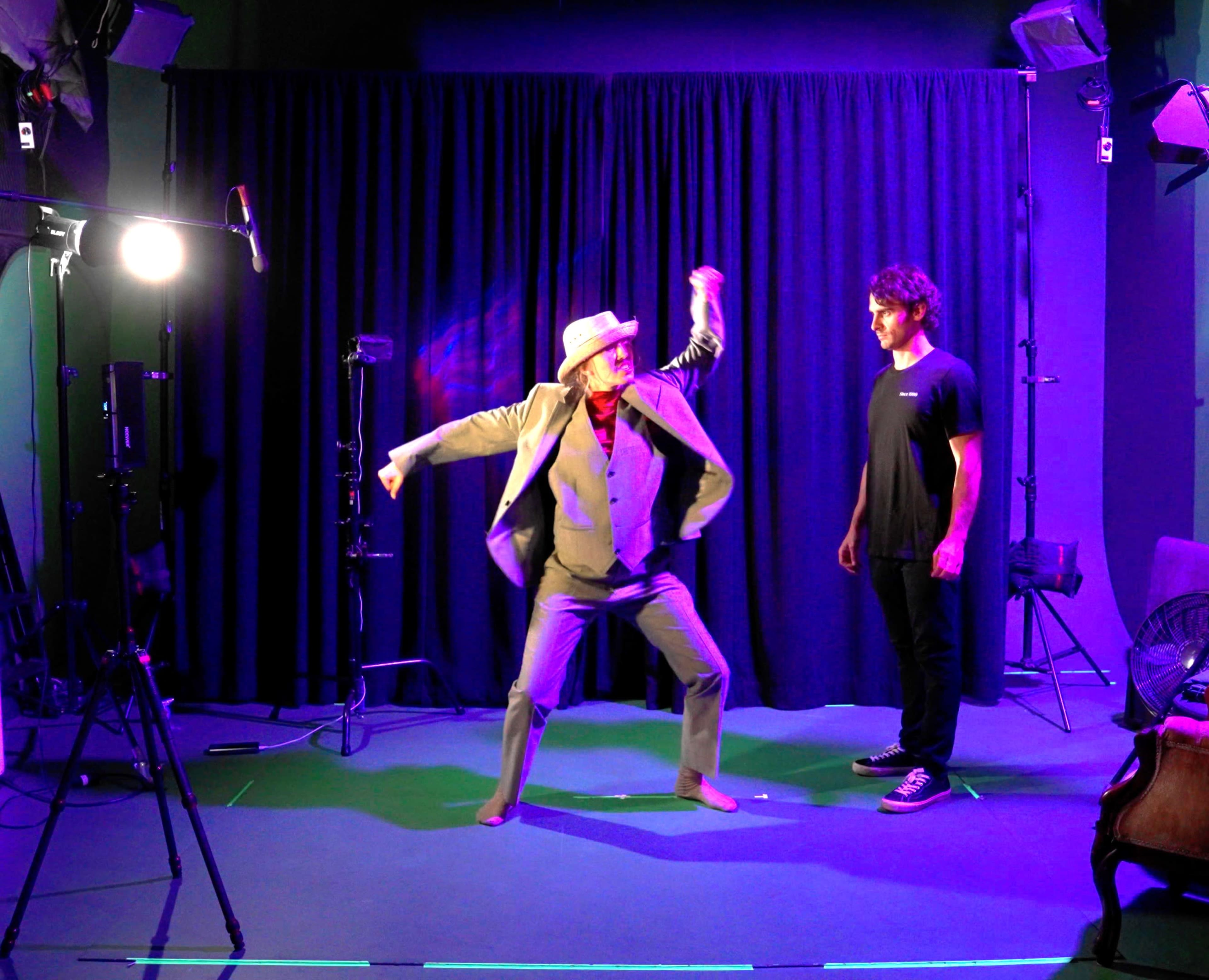 A person in a suit and hat is dancing in front of a man standing on the side, surrounded by studio lights and a dark backdrop.