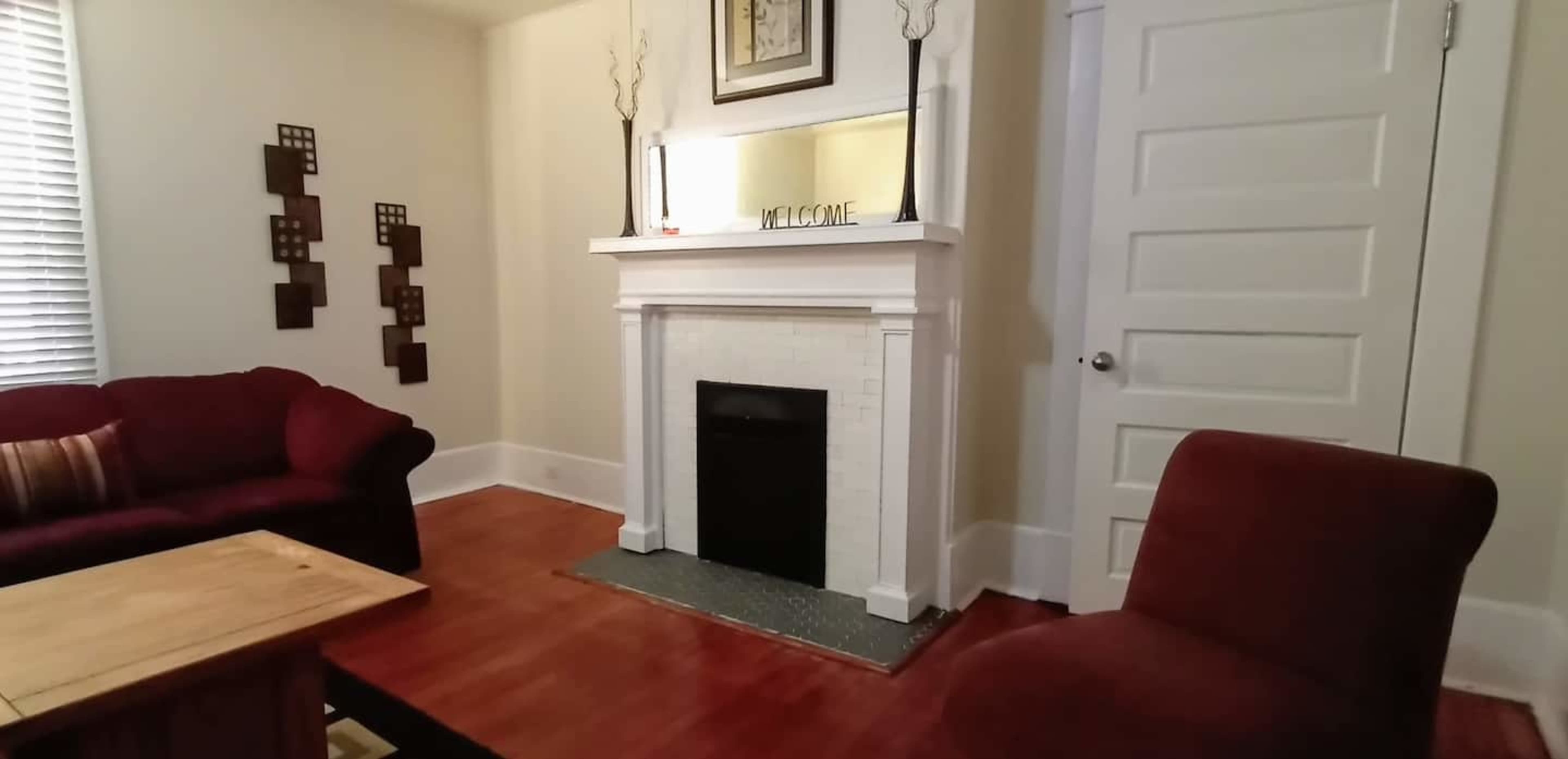 A living room featuring a red sofa, a red armchair, a wooden coffee table, and a fireplace with a mirror above it.