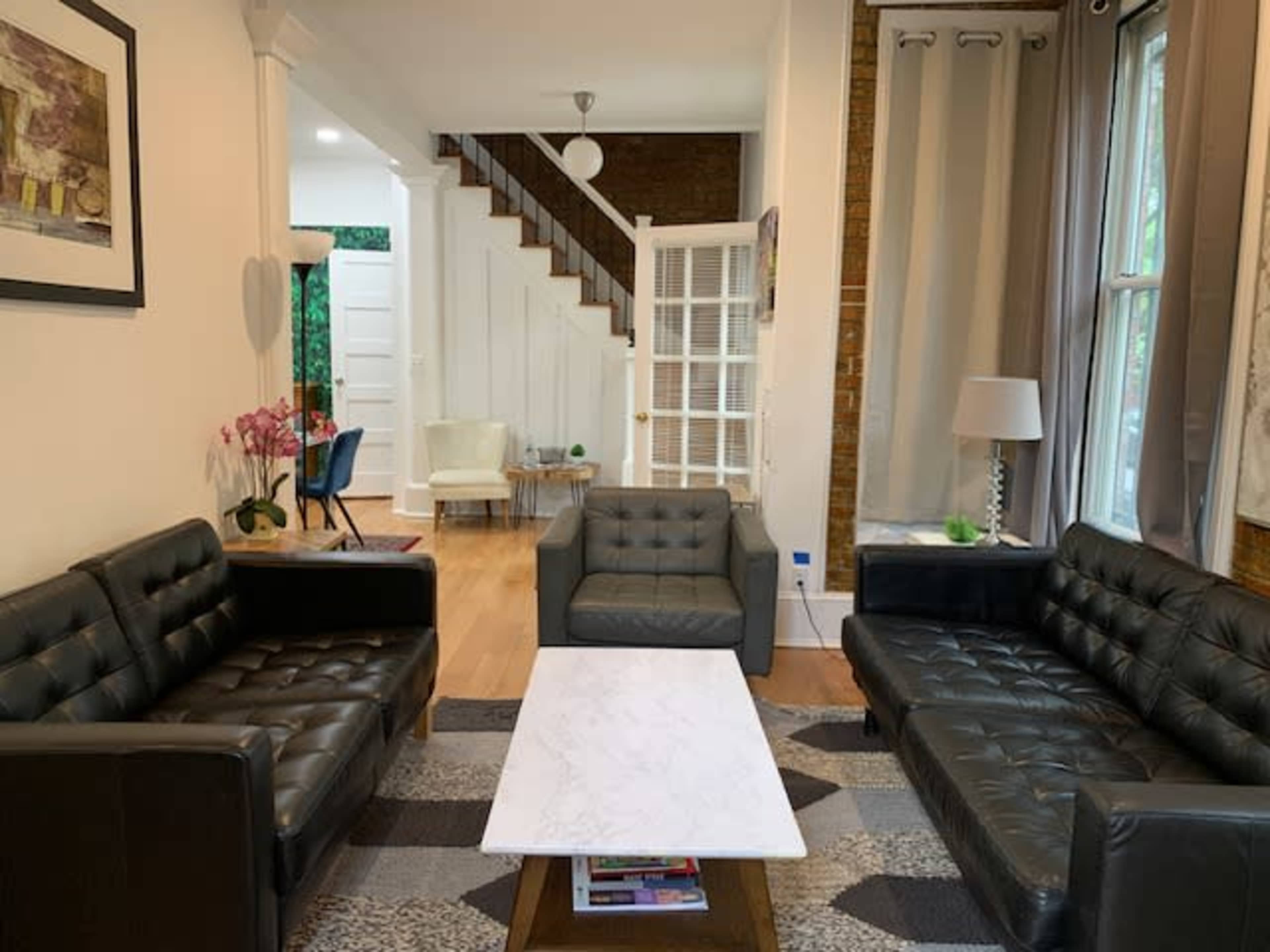 A living room with two black leather sofas facing a marble coffee table, featuring a staircase in the background and a light-filled space with decorative elements.