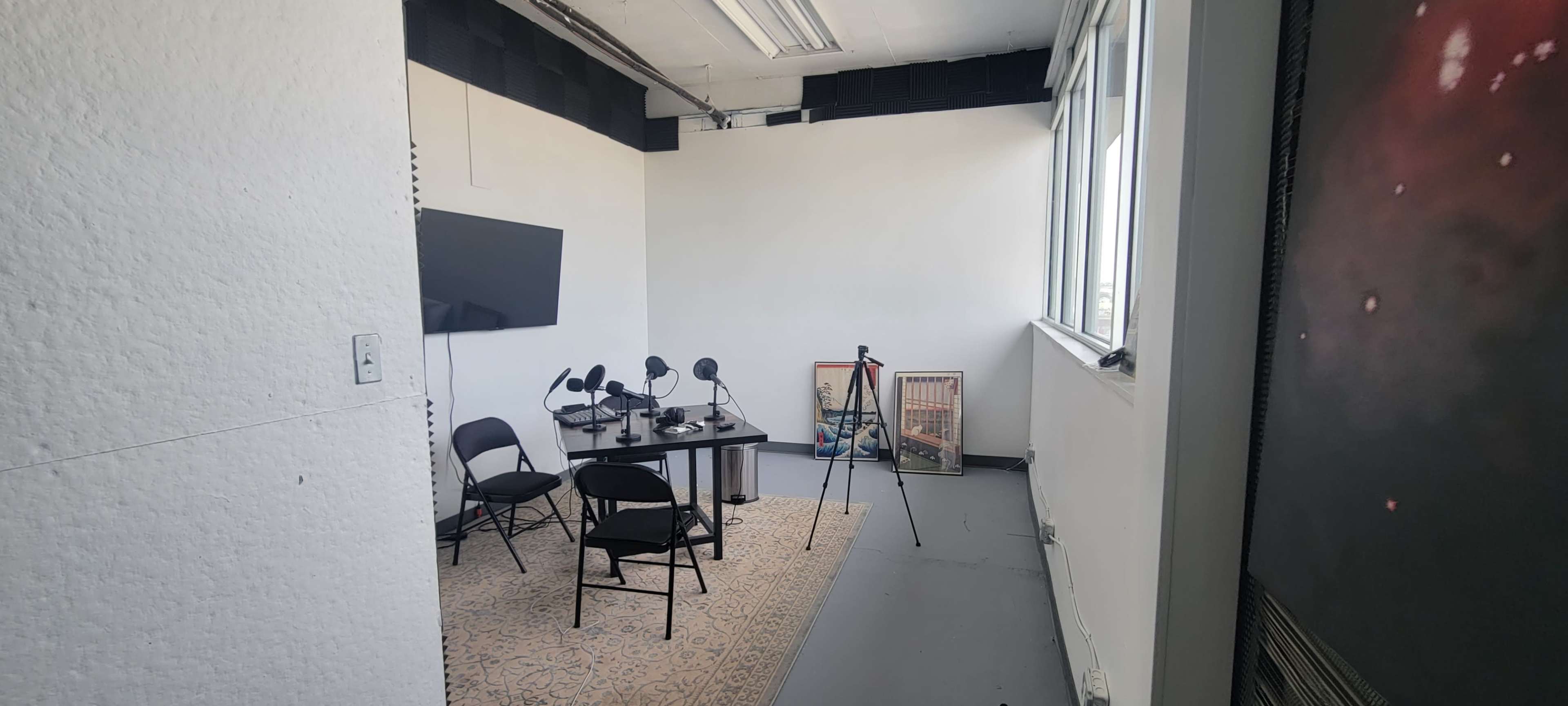A spacious room with a table, four chairs, and microphones, equipped for recording, along with a large screen on the wall.