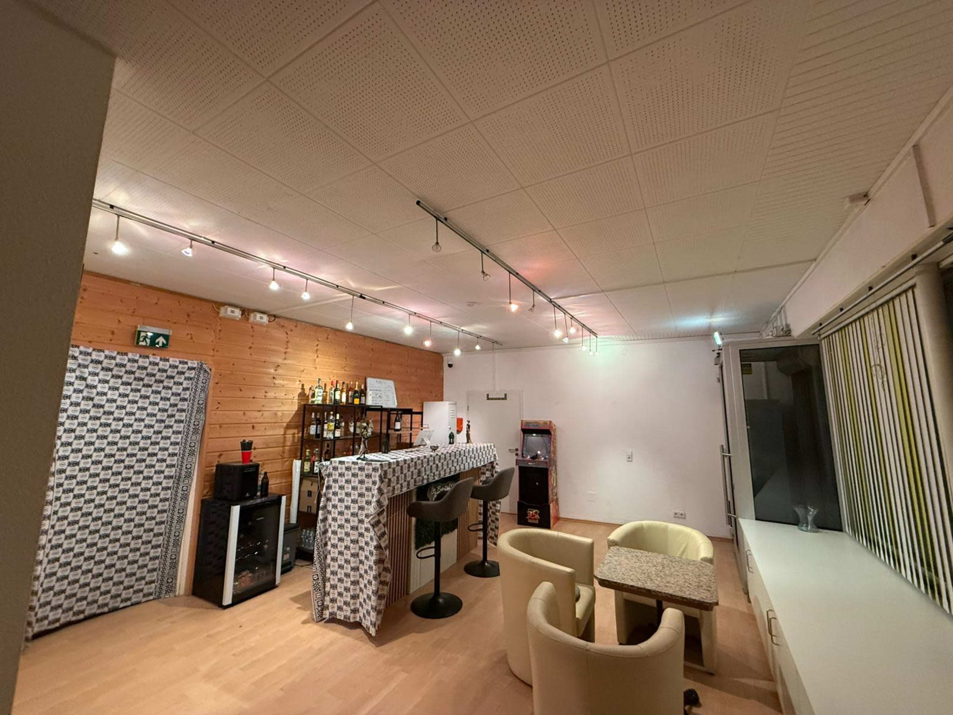 The image shows a spacious, well-lit room set up with a bar area, seating, and shelves displaying various bottles.