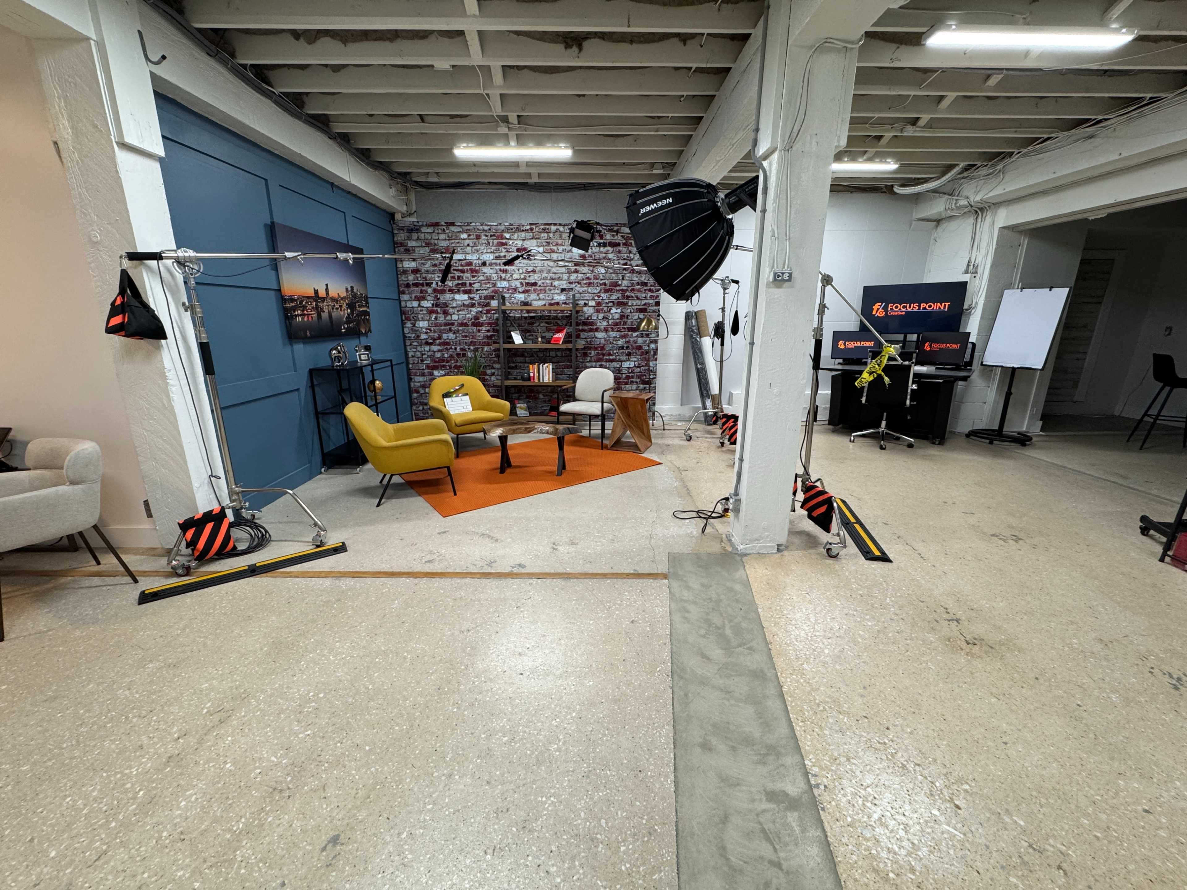 The image shows a studio setup with two chairs, a small table, and various lighting equipment against a backdrop of a brick wall, with additional gear and props visible in the space.