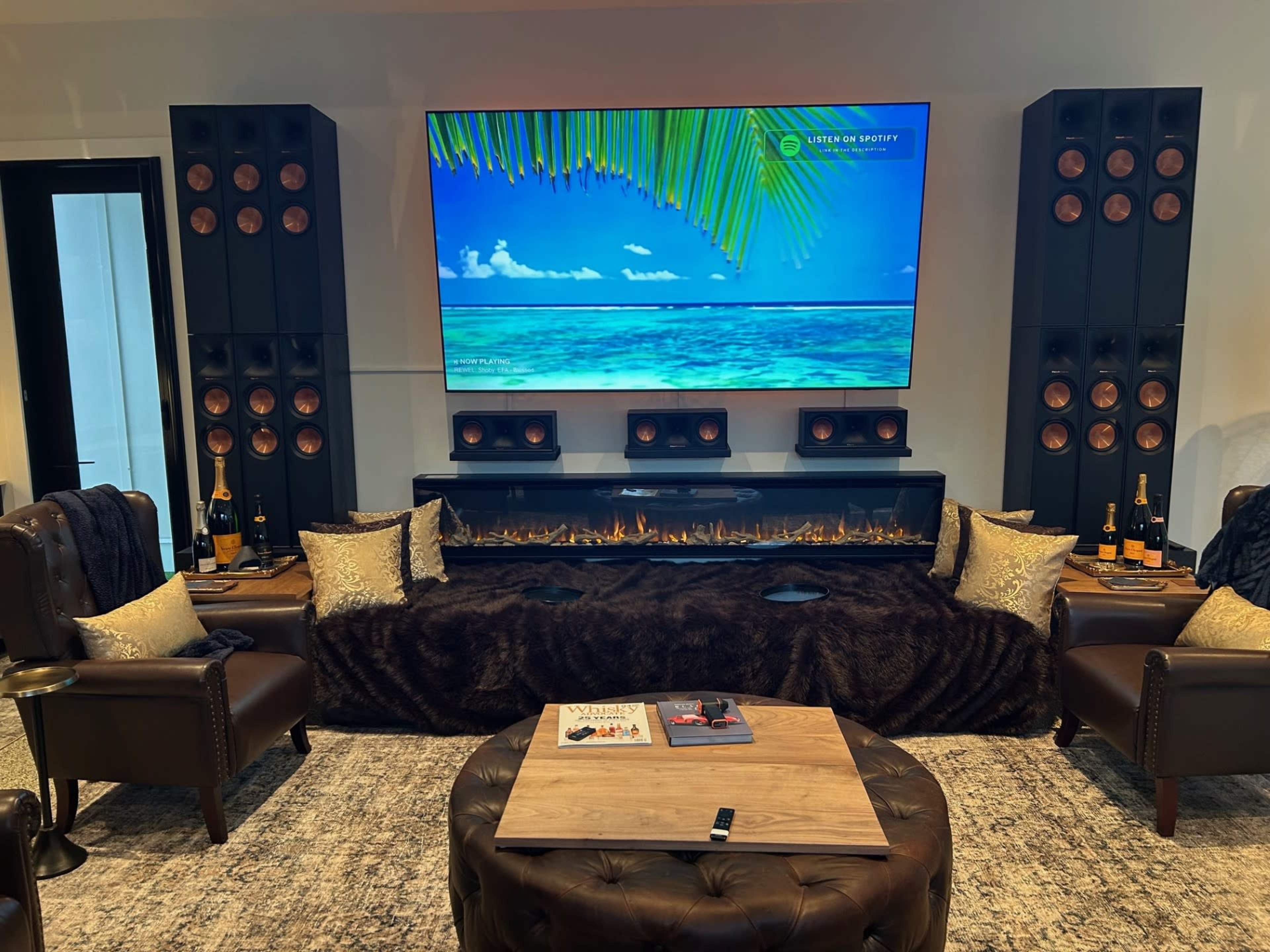 The image shows a modern entertainment room featuring a large television mounted on a wall, flanked by multiple speakers, with plush seating, a coffee table, and decorative items including champagne bottles and pillows.