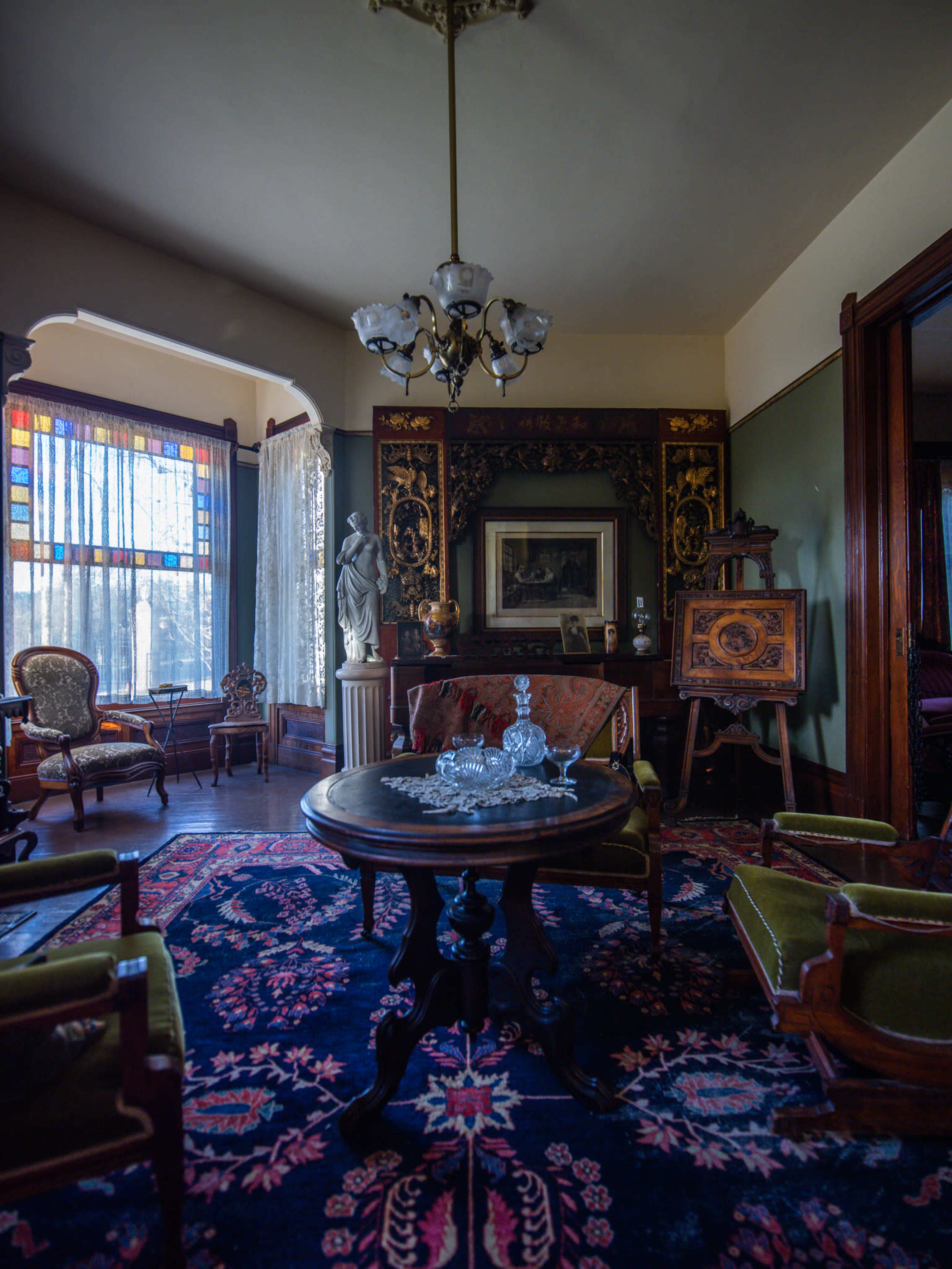 A vintage parlor with ornate furniture, a patterned rug, and stained glass windows illuminating the space.