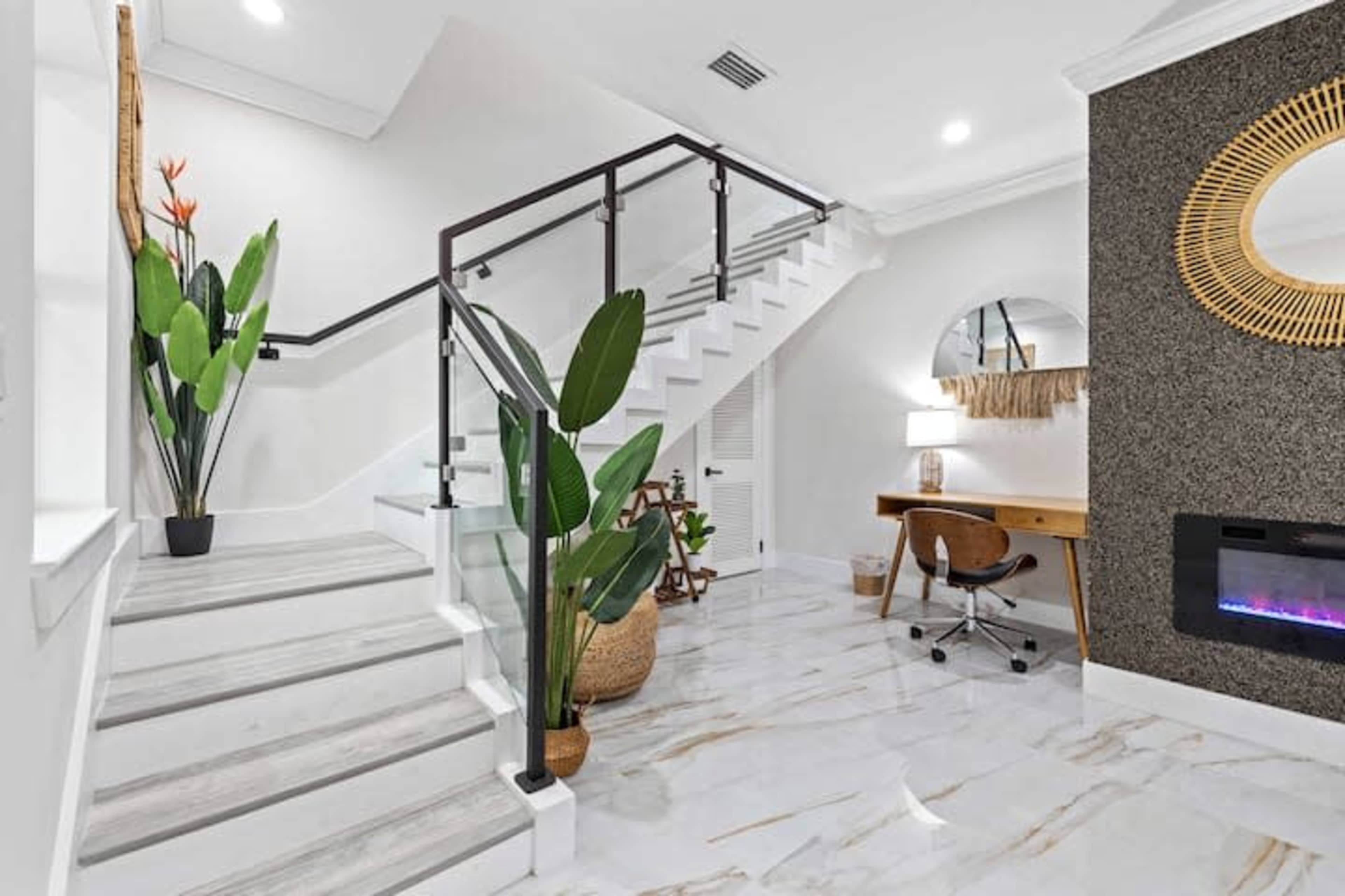 A modern staircase with glass railings, surrounded by decorative plants and a small desk area, all set in a room with marble flooring.