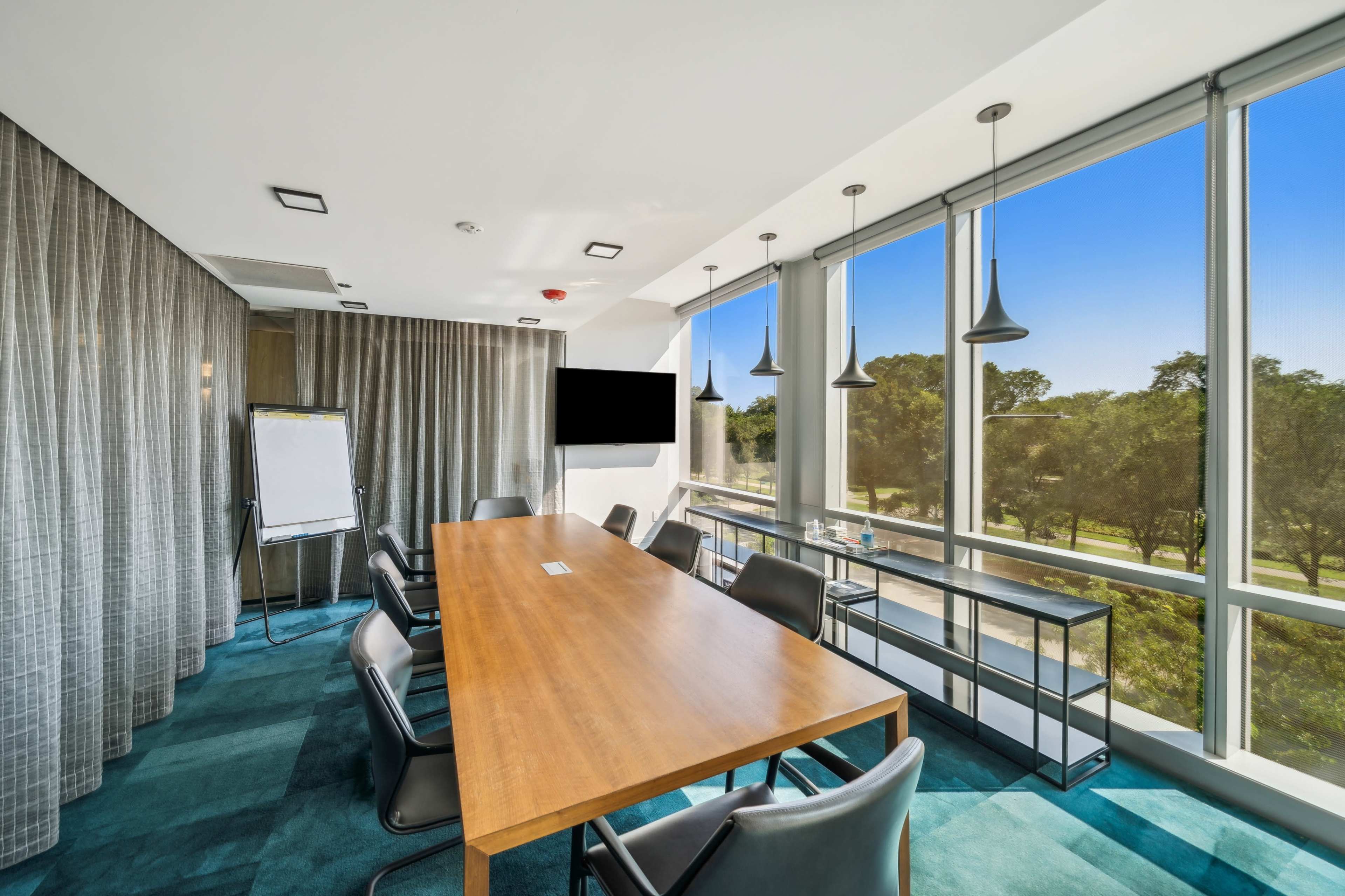A modern meeting room features a long wooden table surrounded by black chairs, large windows with a view of greenery, and a presentation screen.