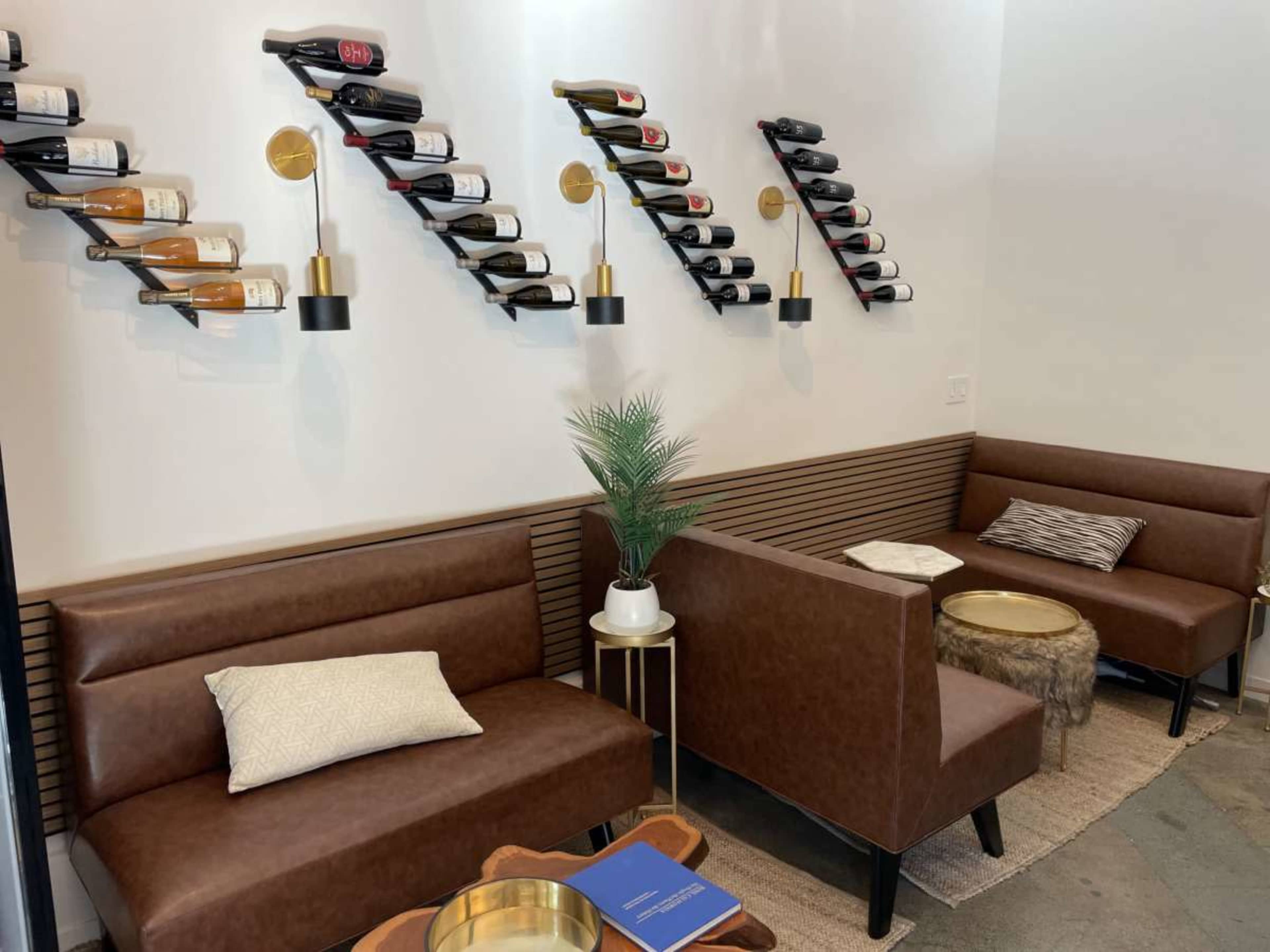The image shows a modern seating area with two brown upholstered couches, two small tables, a decorative plant, and wall-mounted wine racks displaying several bottles of wine.