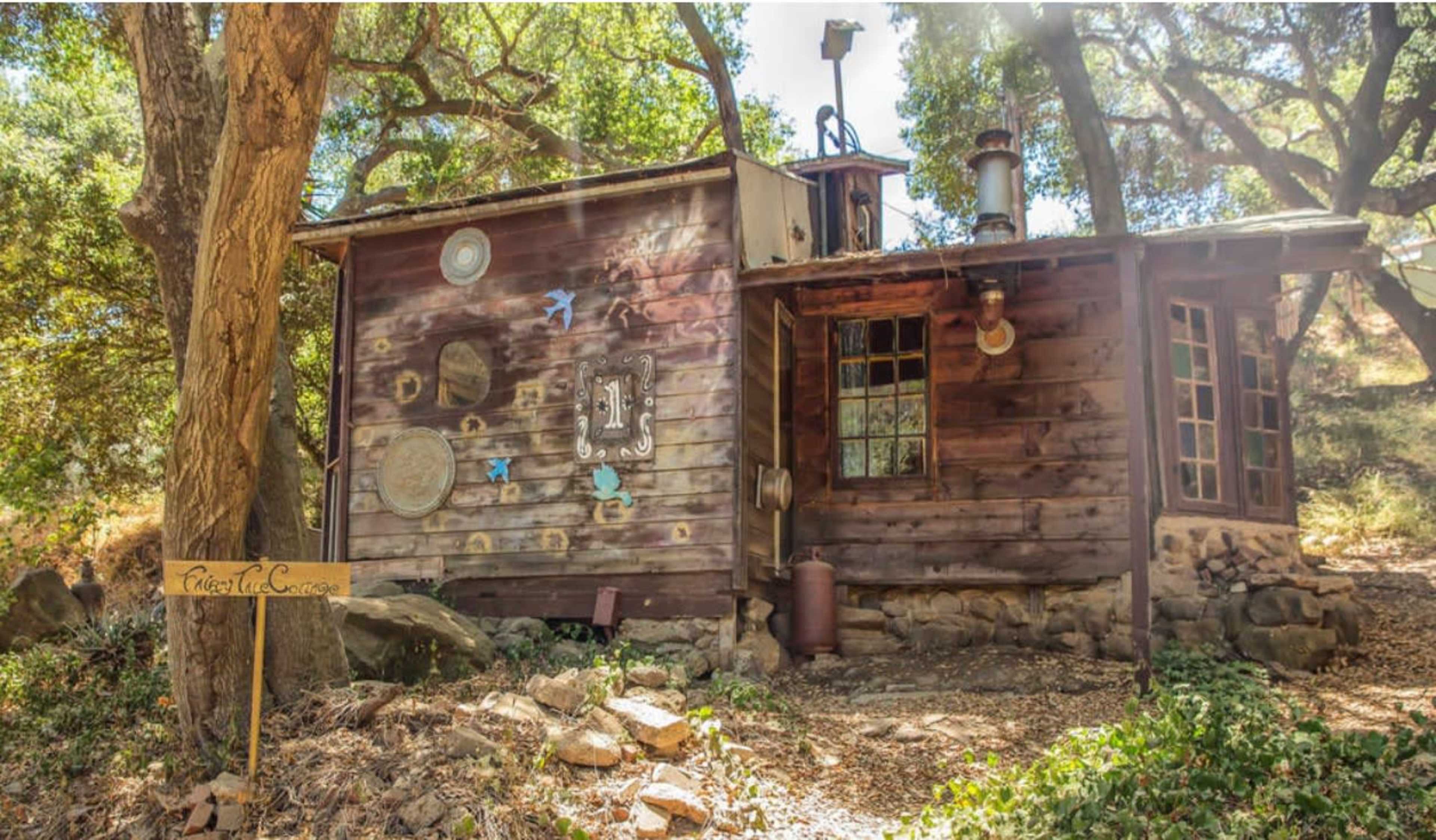 A small, rustic wooden cabin with a stone foundation, set among trees in a natural landscape.