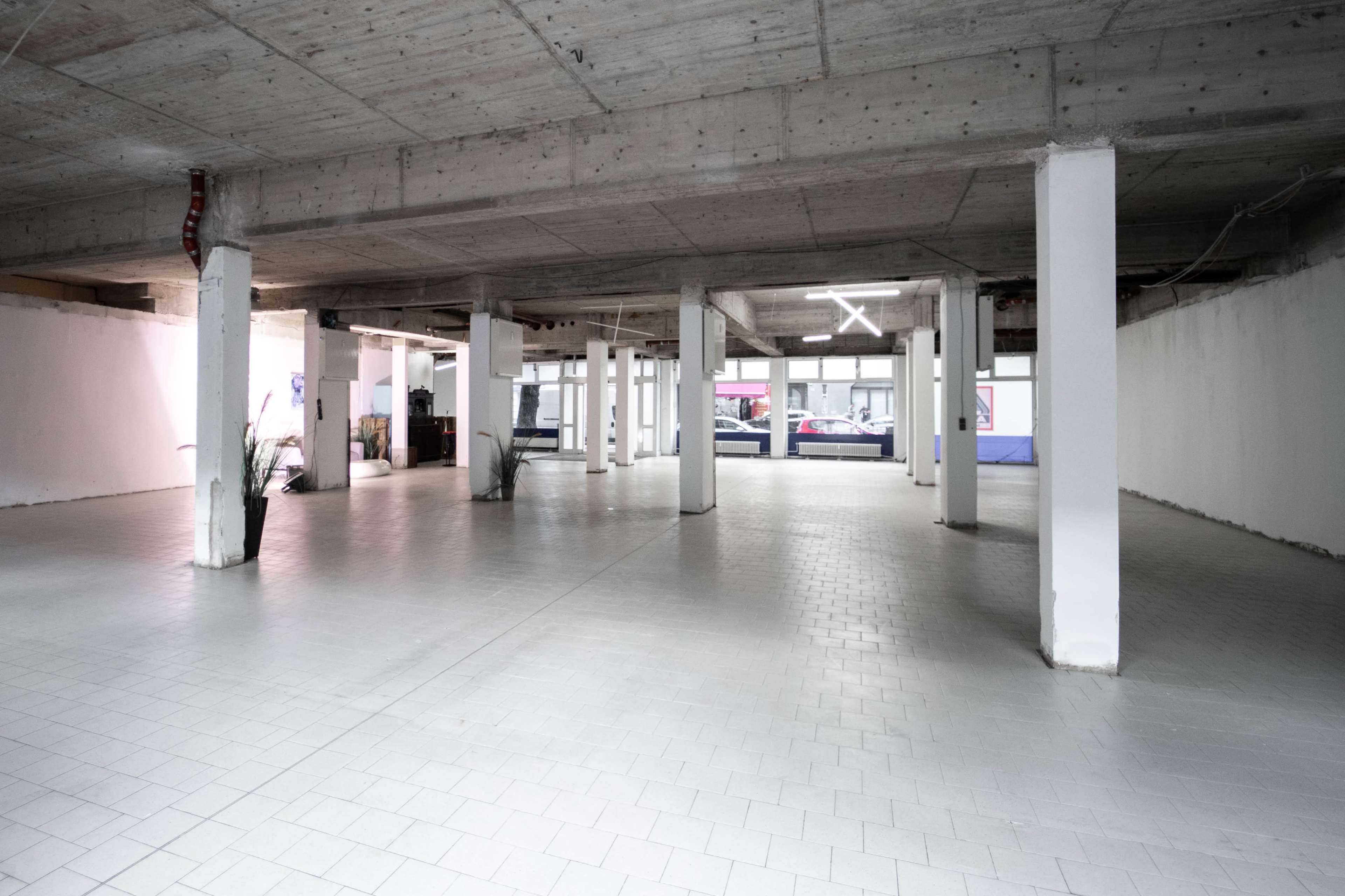 A spacious, empty room with concrete pillars, tiled flooring, and large windows along one wall.