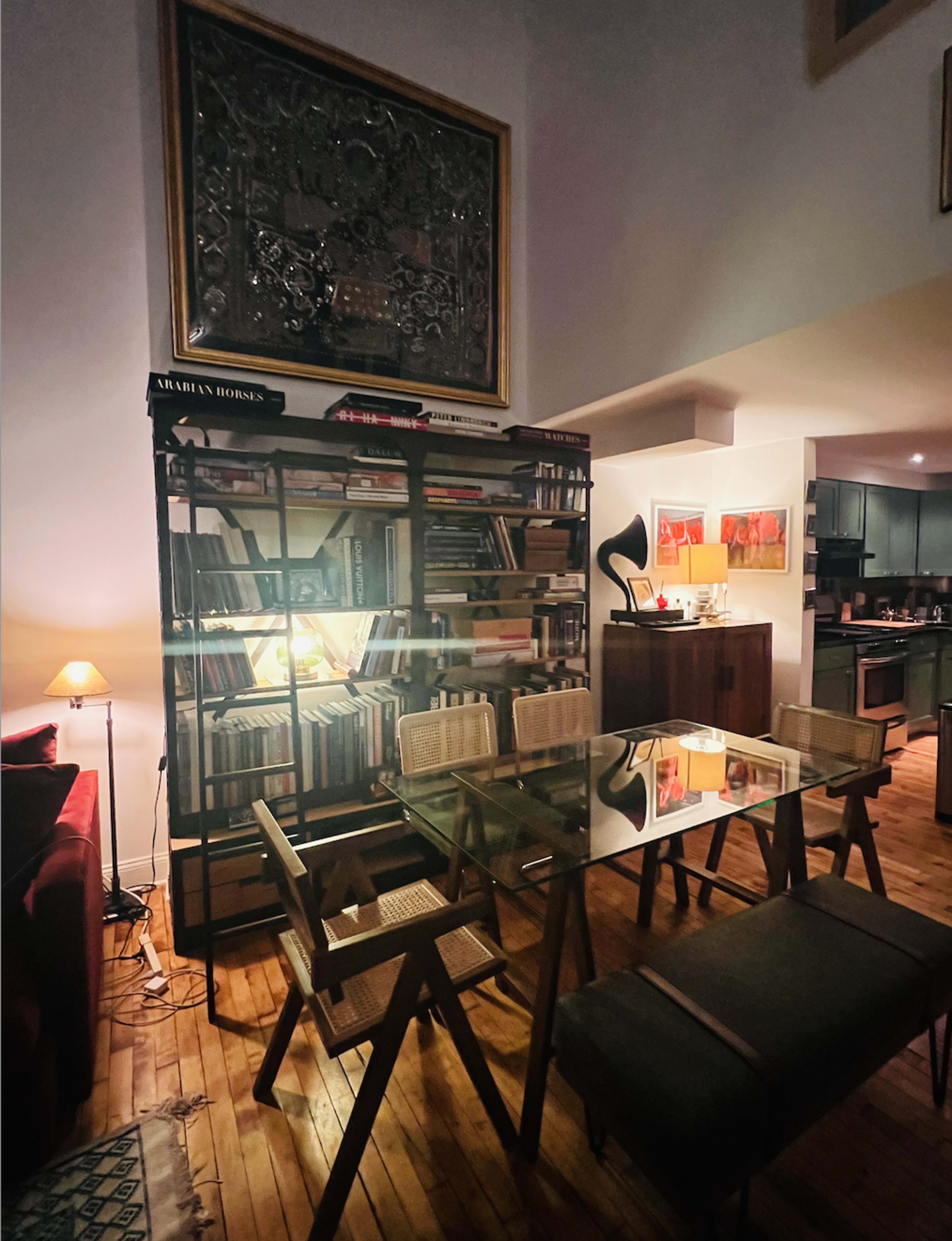 A cozy dining area with a glass table, surrounded by wooden chairs, and a bookshelf filled with books, set against a softly lit backdrop.
