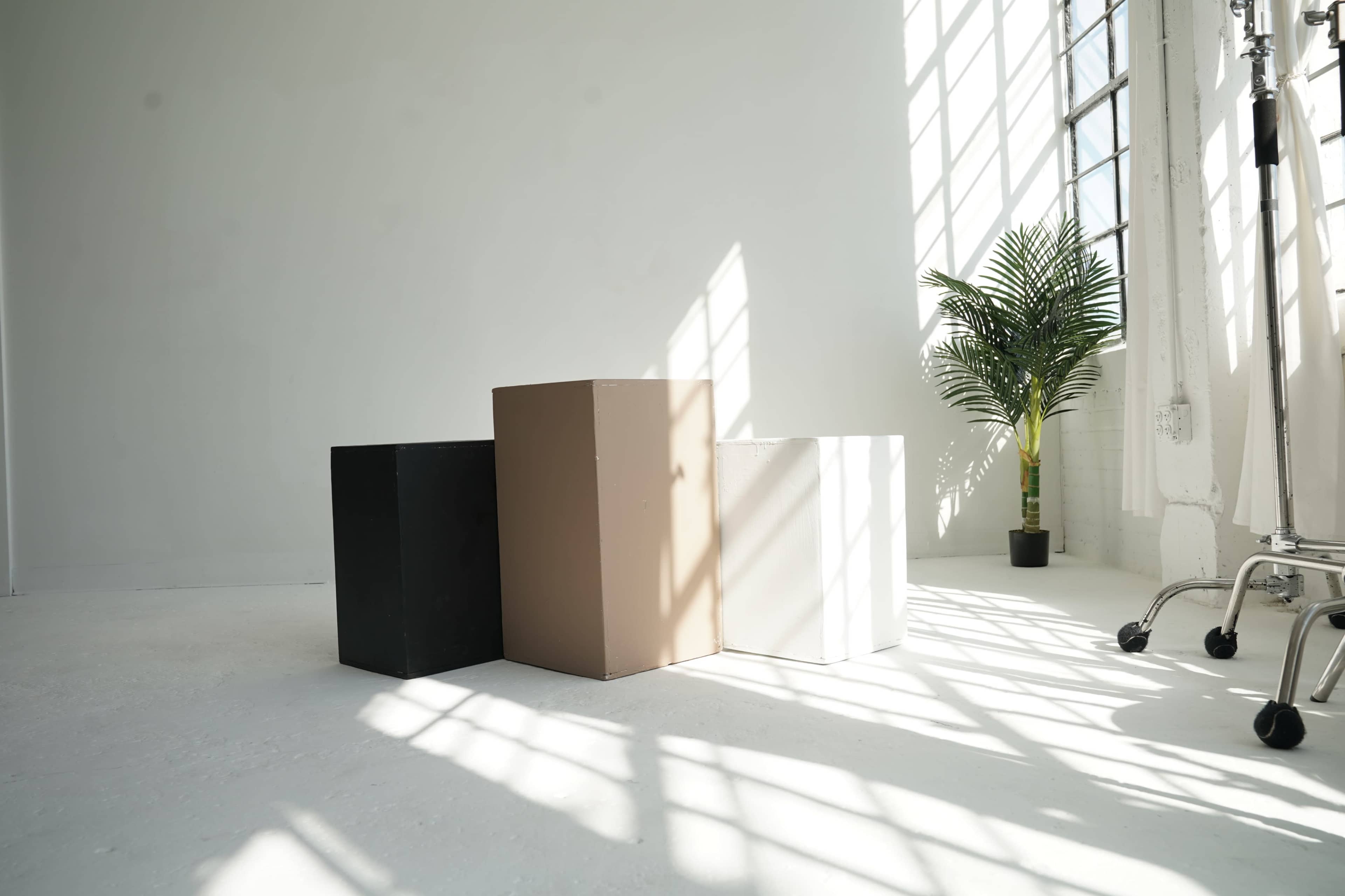 The image shows three geometric boxes in black, beige, and white arranged on a light-colored floor, with large windows casting shadows and a potted plant in the background.
