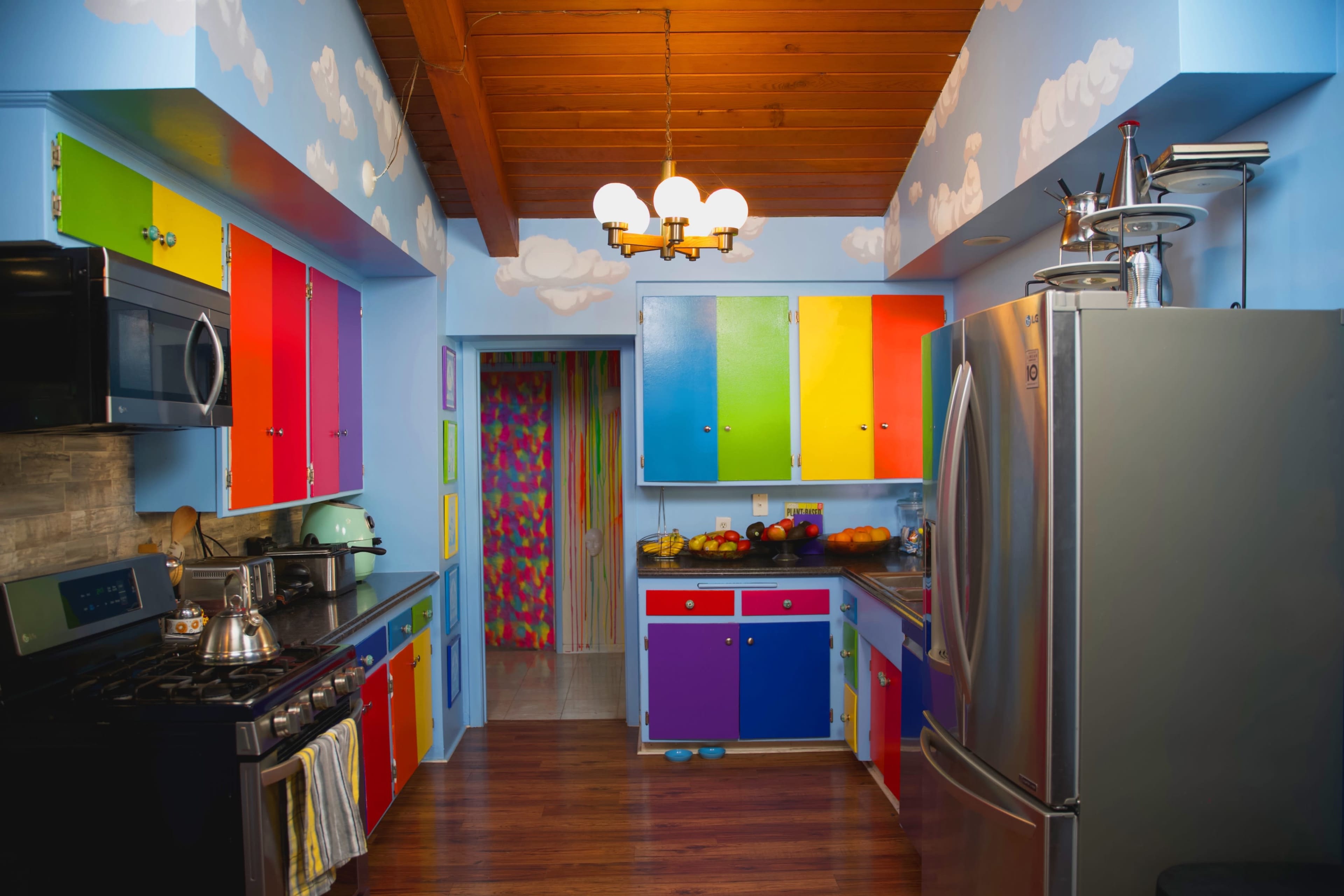 A colorful kitchen featuring bright cabinets, a stainless steel refrigerator, and a wooden ceiling with painted clouds on the walls.
