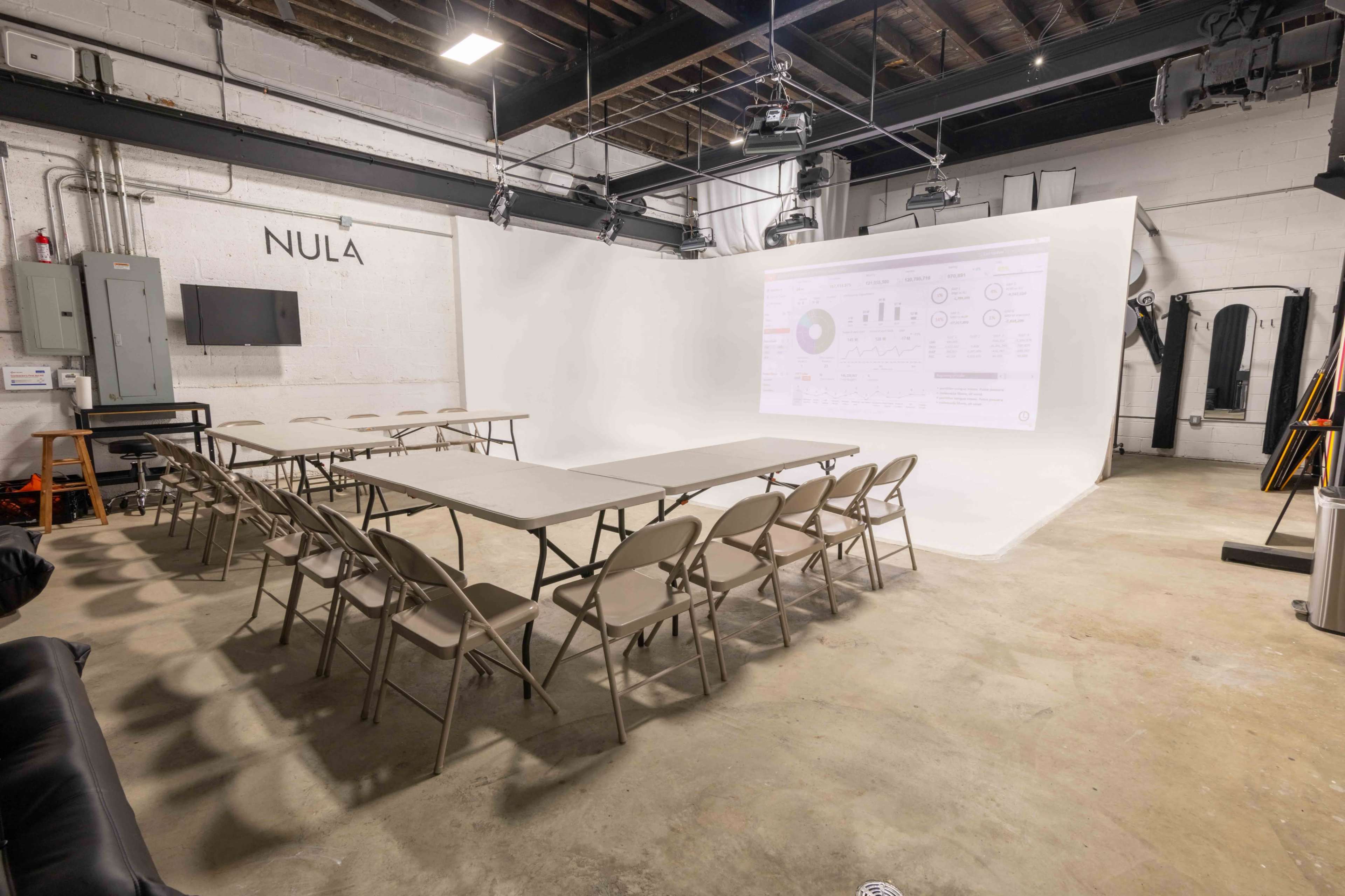 The image shows a spacious room set up for a meeting, featuring a white backdrop, multiple folding tables arranged in a U-shape, and chairs positioned around them.
