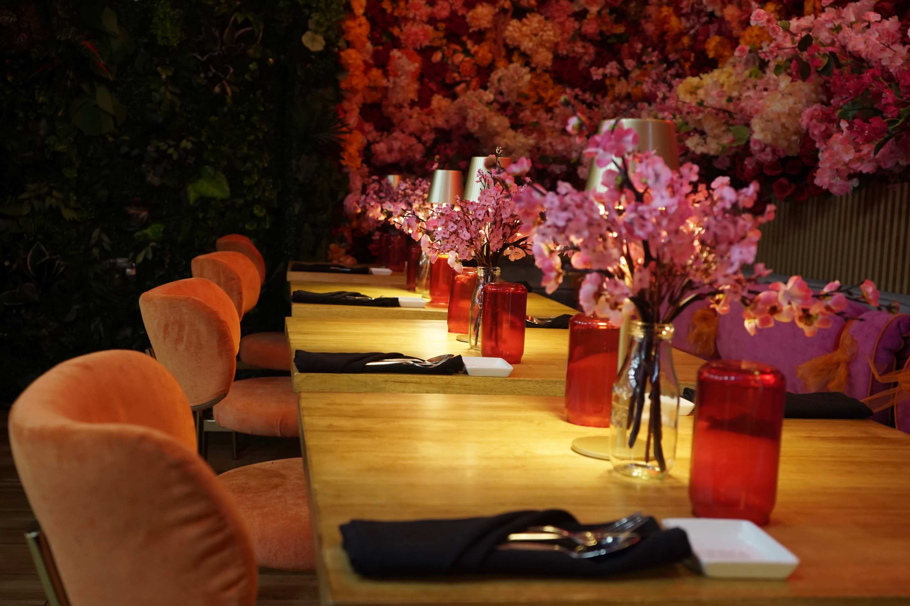 A long wooden dining table is set with red vases of pink flowers and illuminated by table lamps, against a backdrop of a floral wall.