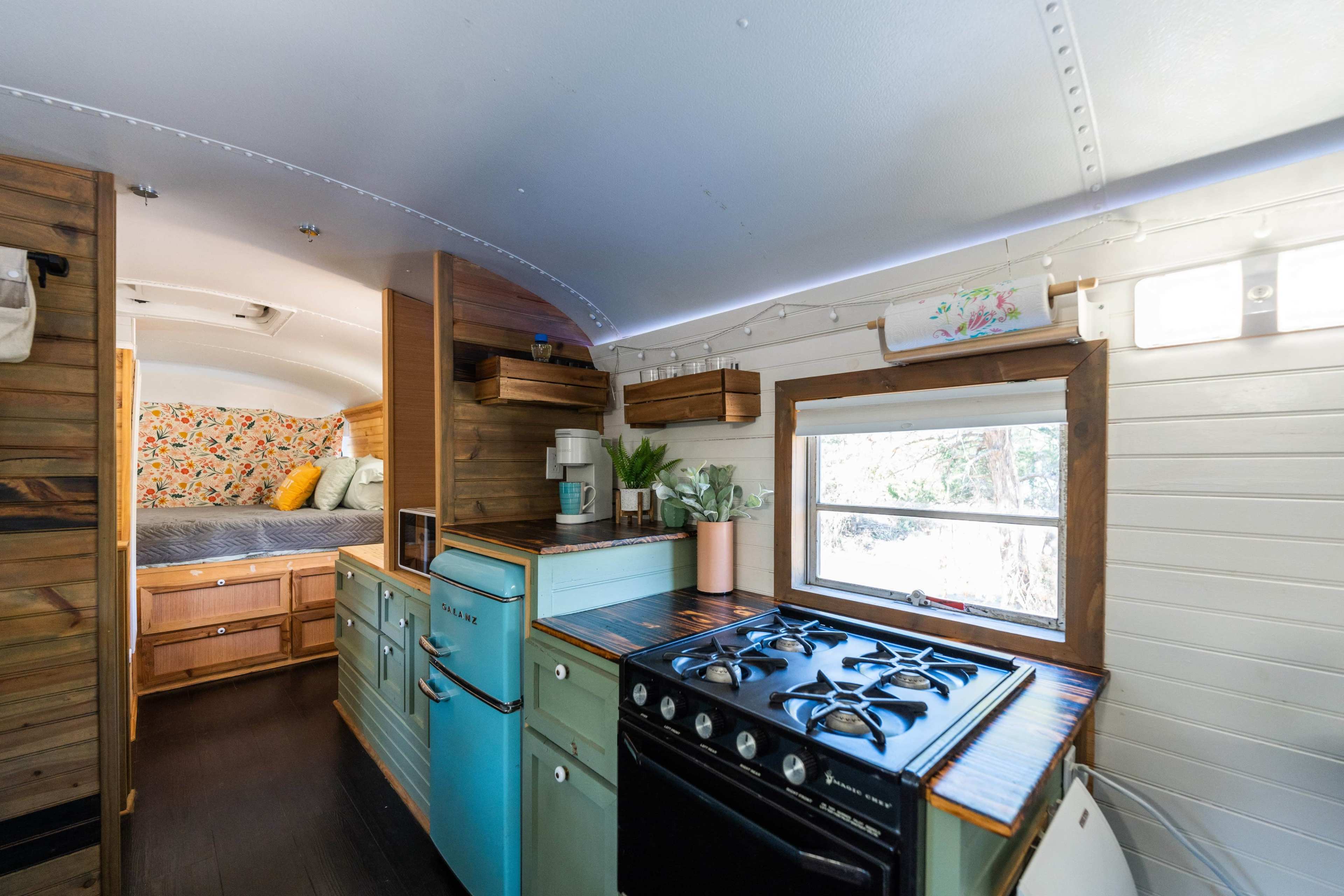 The image shows a compact kitchen inside a converted bus, featuring a gas stove, a sink, and wooden cabinetry with floral decor in the background.
