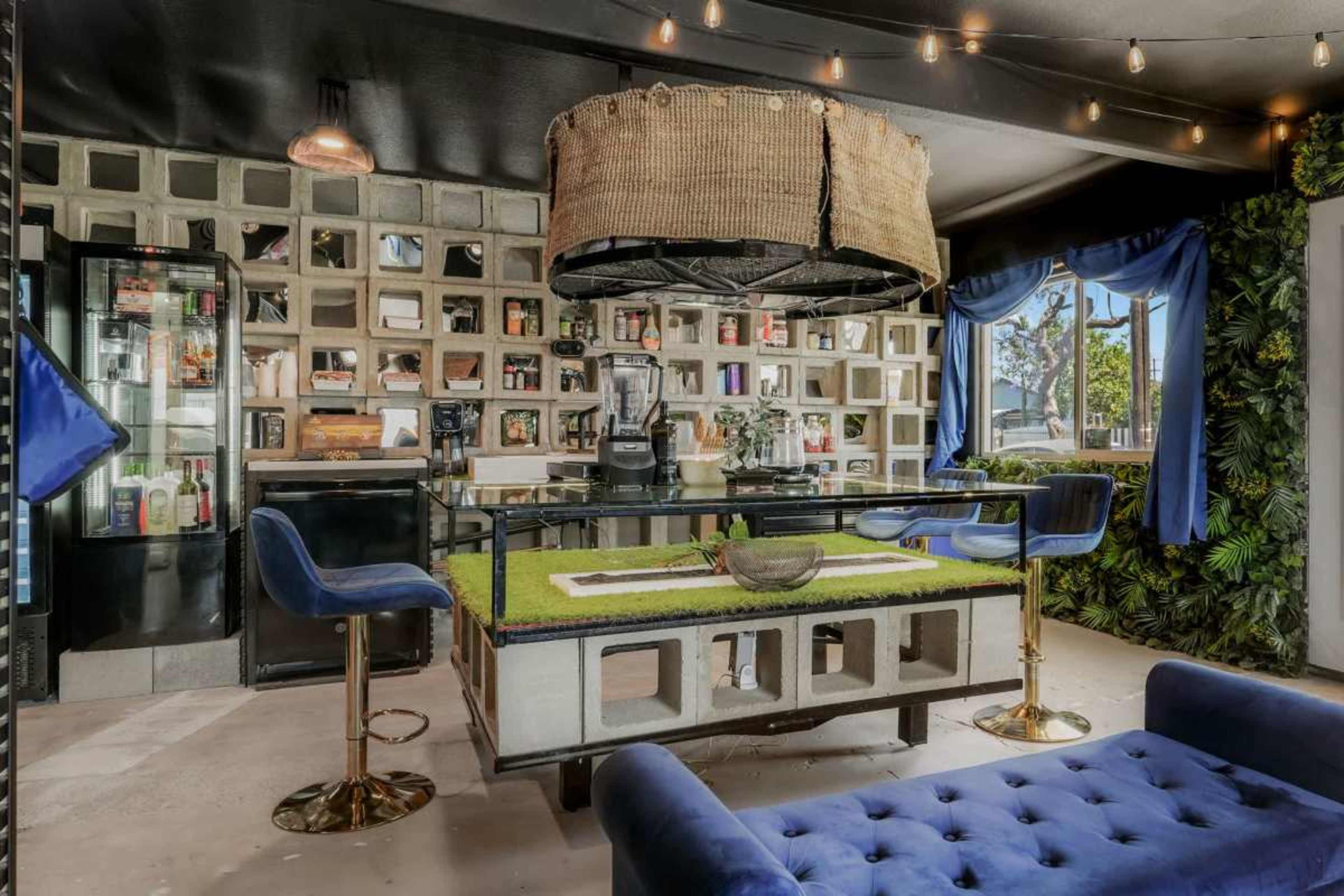 The image shows a modern bar area featuring a green-topped island, a wall of patterned blocks, and a variety of bottles displayed in glass cabinets.