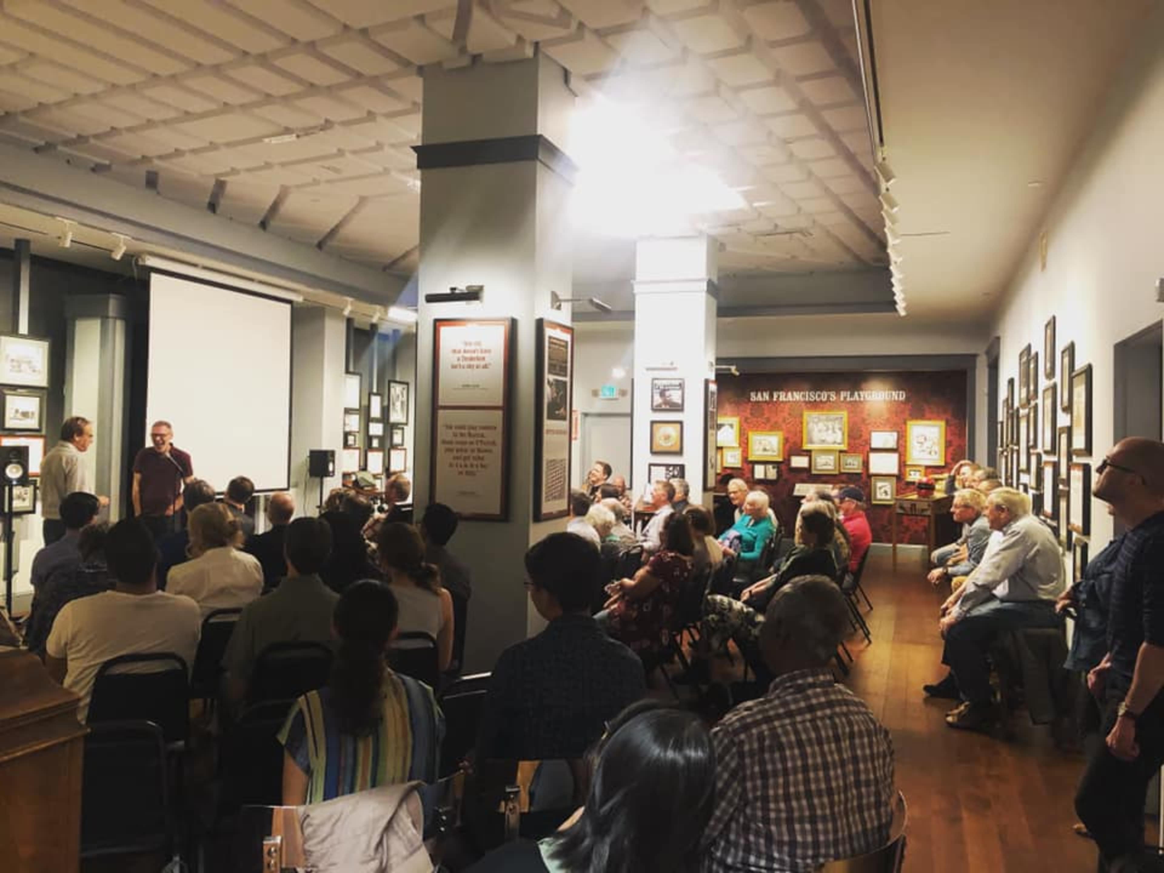 A crowd of people sits facing a speaker in a room adorned with framed photographs and artwork.