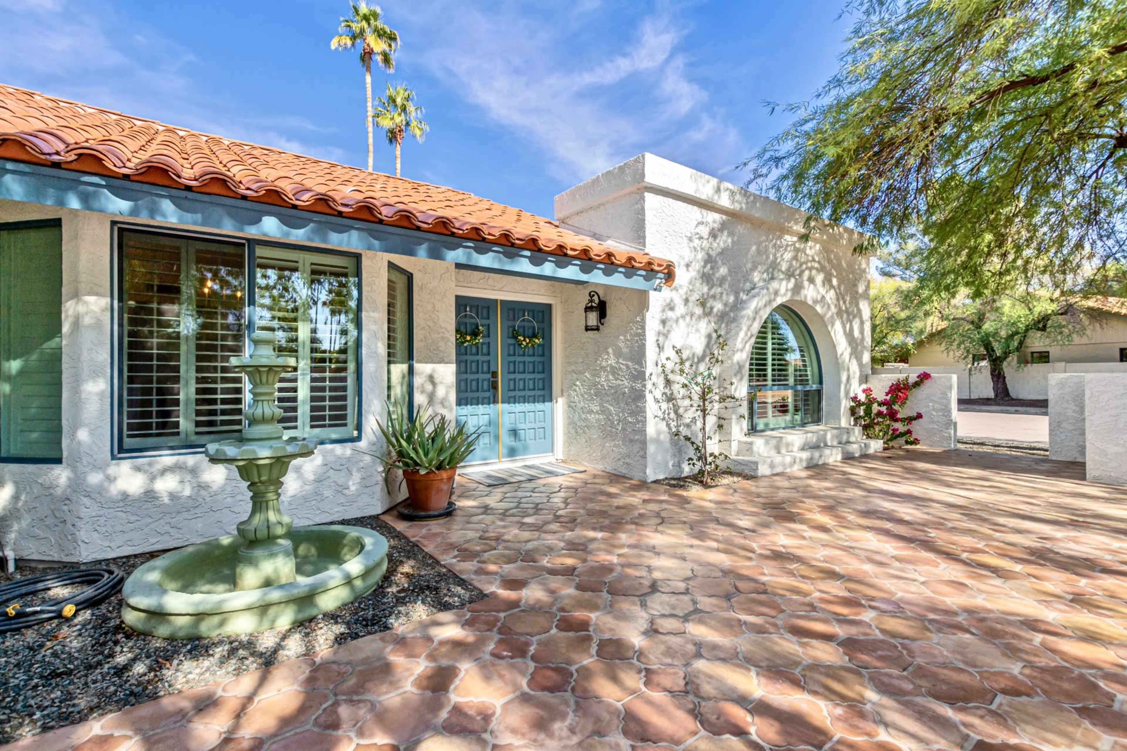 A courtyard features a tiled walkway, a fountain, and potted plants alongside a white stucco building with blue doors and a red tiled roof.