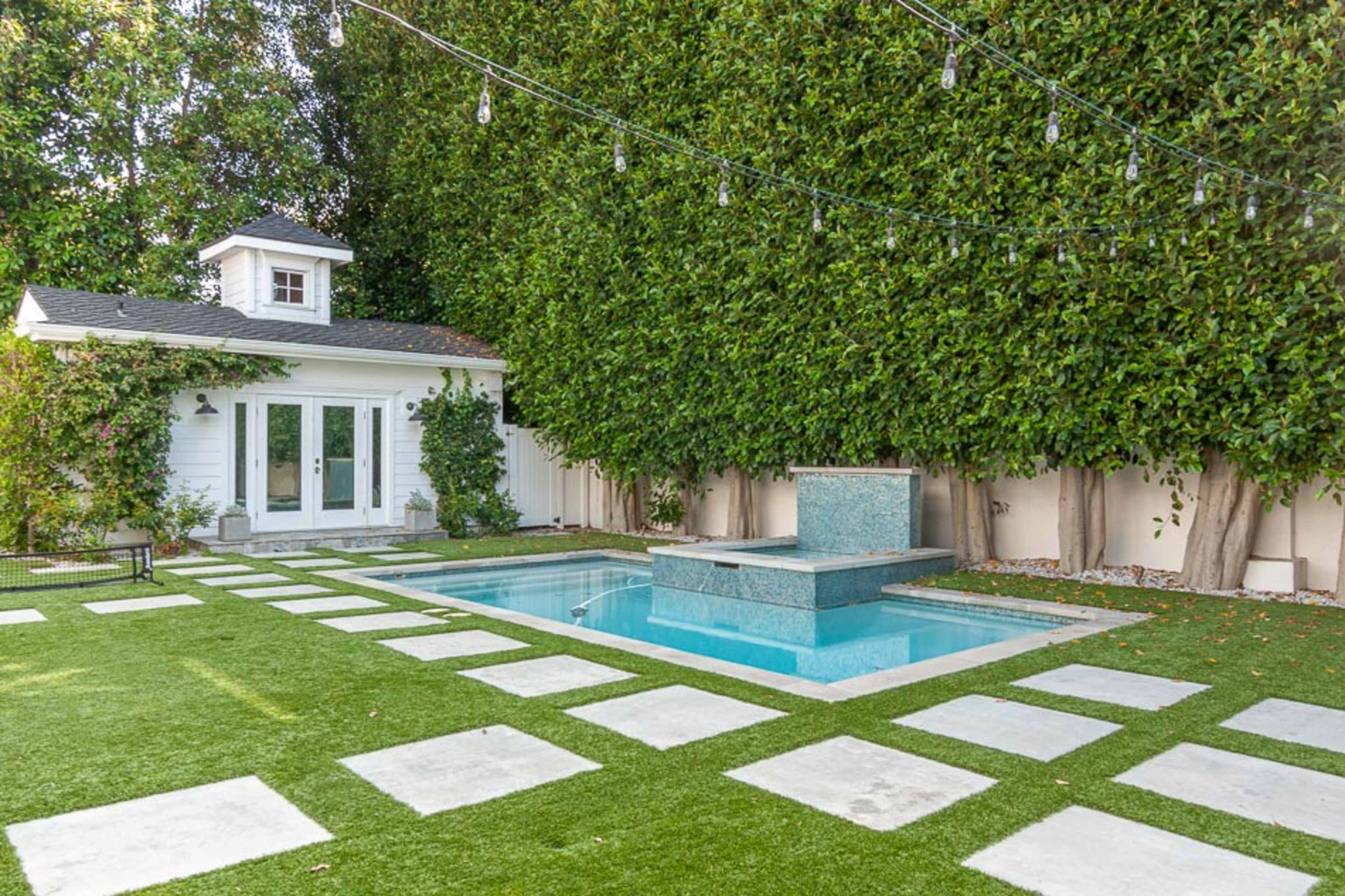 A small swimming pool is surrounded by a neatly manicured lawn and stone pathways, next to a white shed with a cupola and greenery along the fence.
