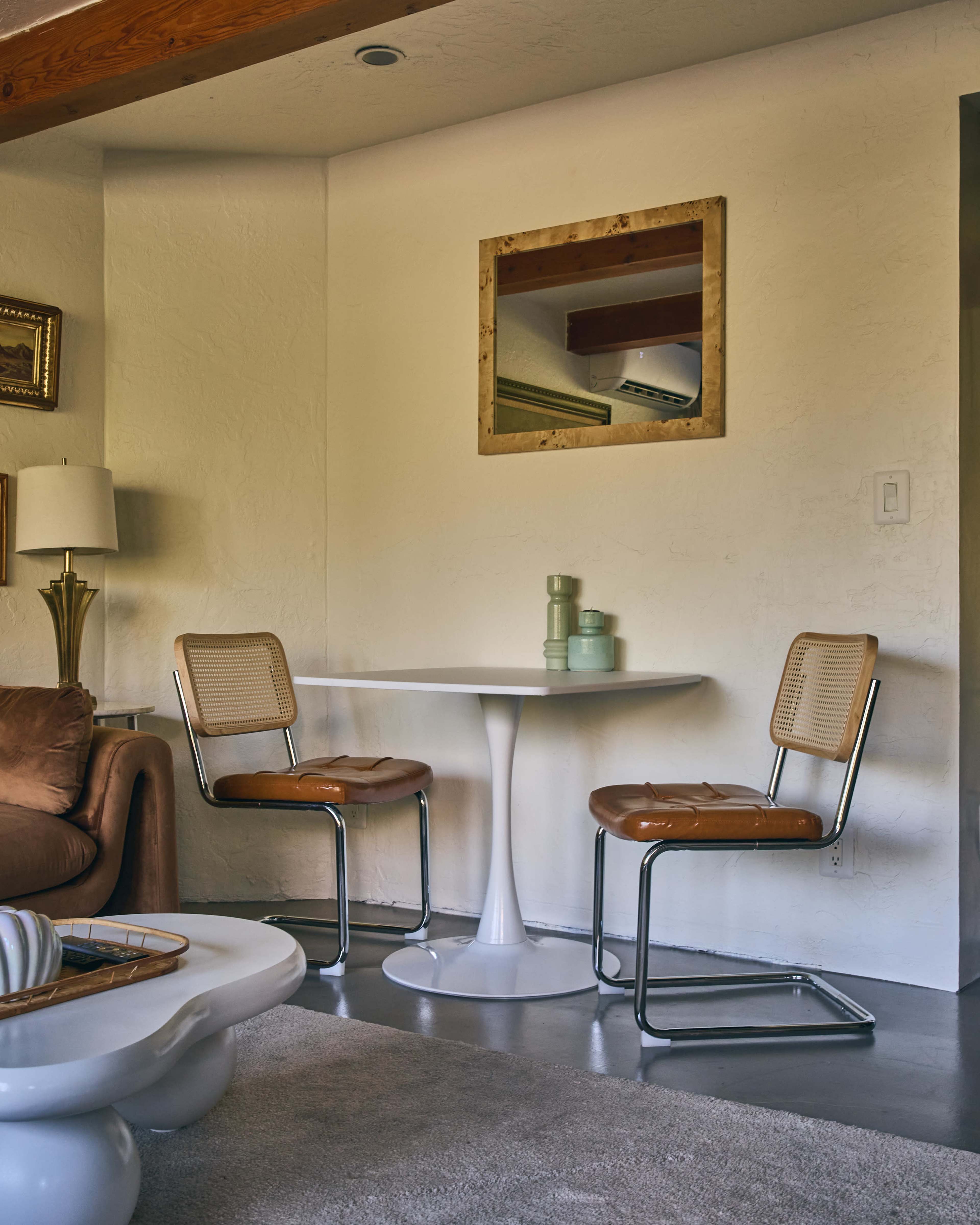 A small dining area features a white table with two modern chairs against a textured wall, accompanied by a lamp and a framed mirror.