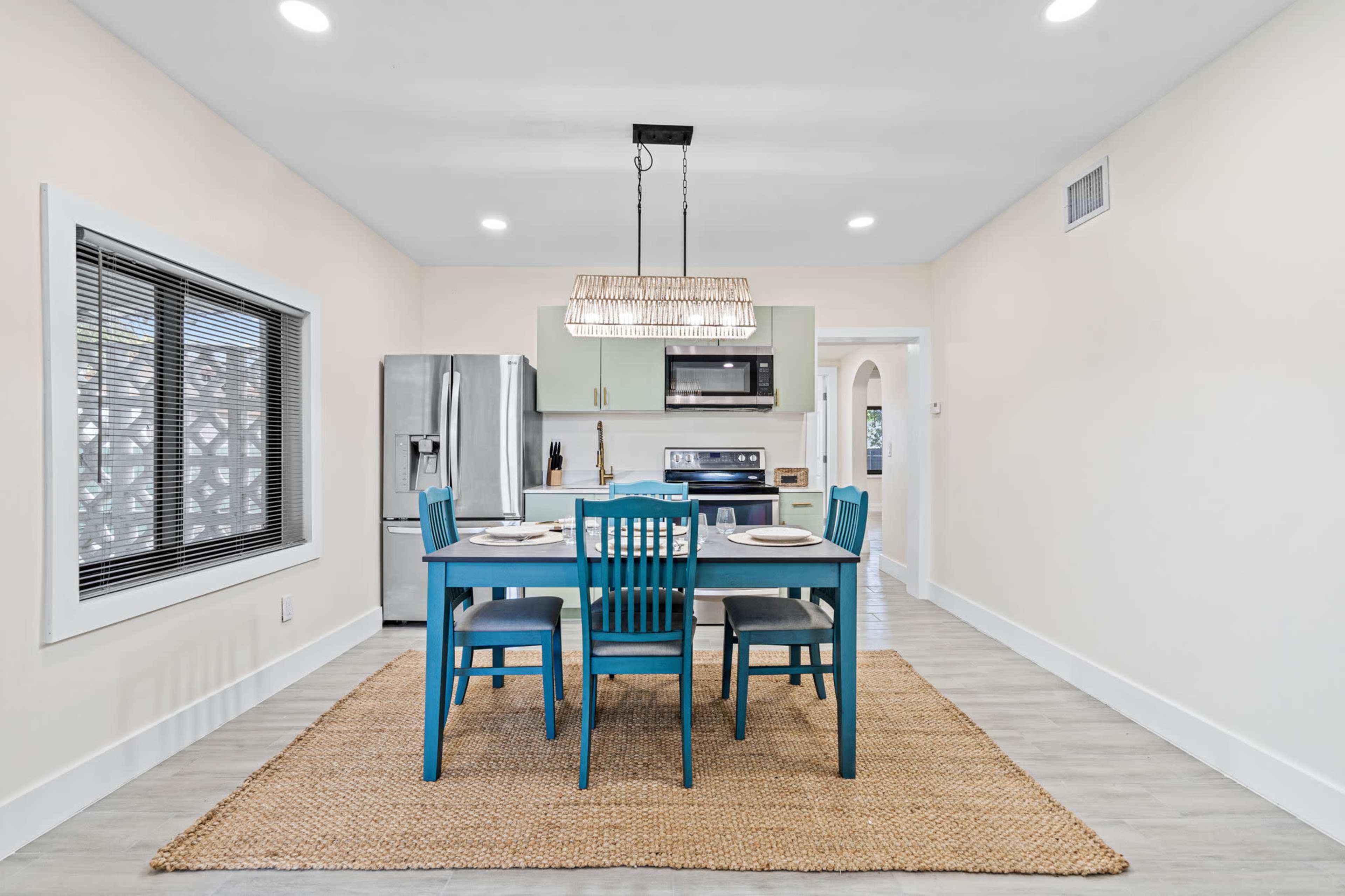 The image depicts a modern kitchen with a dining area featuring a rectangular table and blue chairs, placed on a woven rug, with stainless steel appliances in the background.