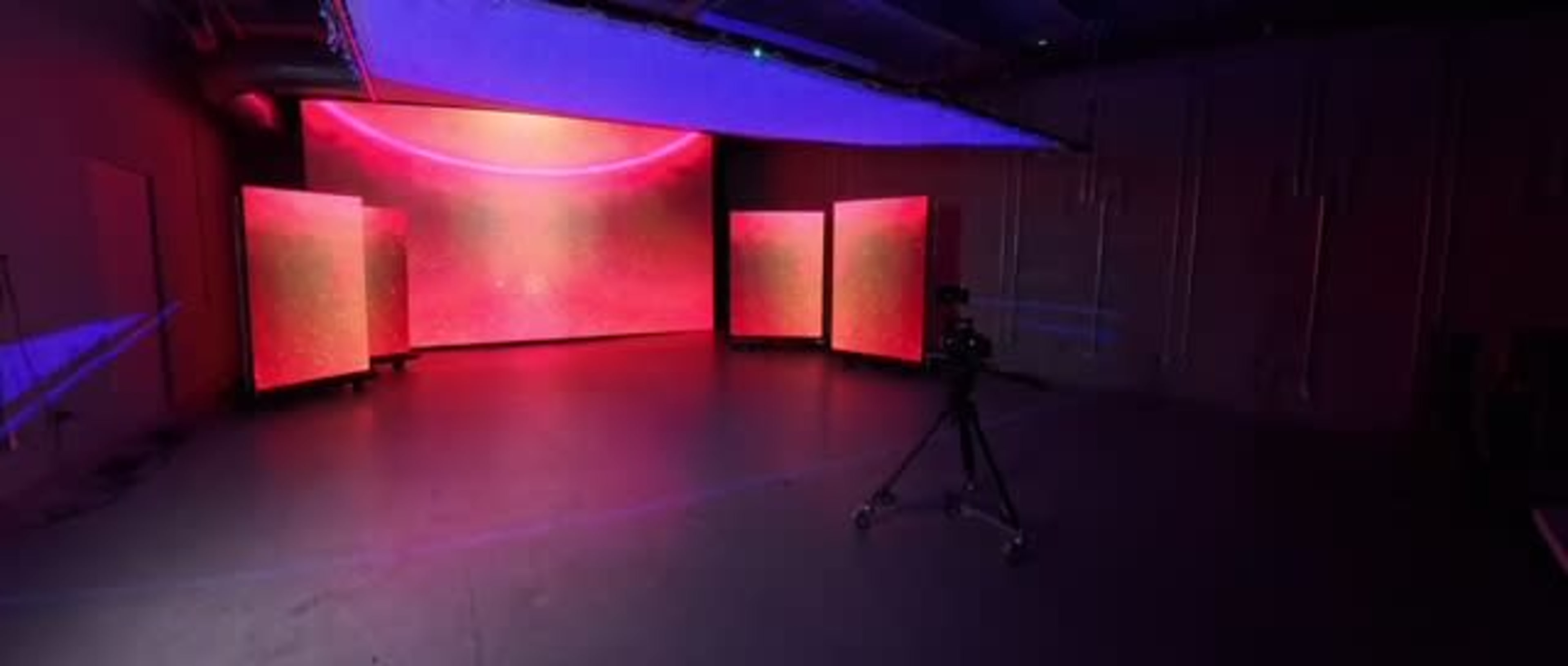 The image shows a studio space with a large, illuminated backdrop featuring gradient colors and three illuminated panels, along with a camera on a tripod positioned in front of them.