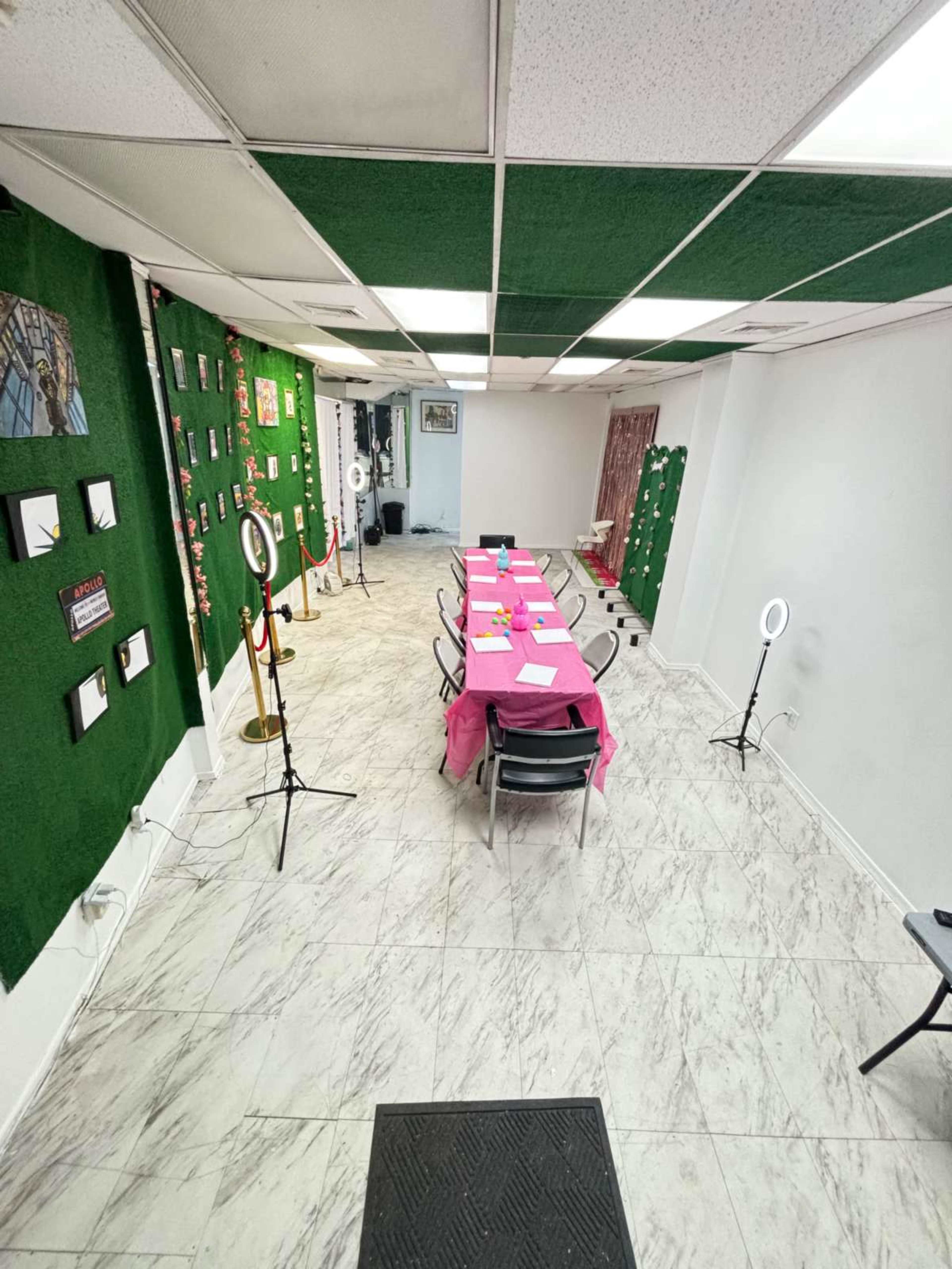 A spacious room set up for an event, featuring a long table covered with a pink tablecloth, surrounded by chairs, and decorated walls with photo displays and lighting equipment.