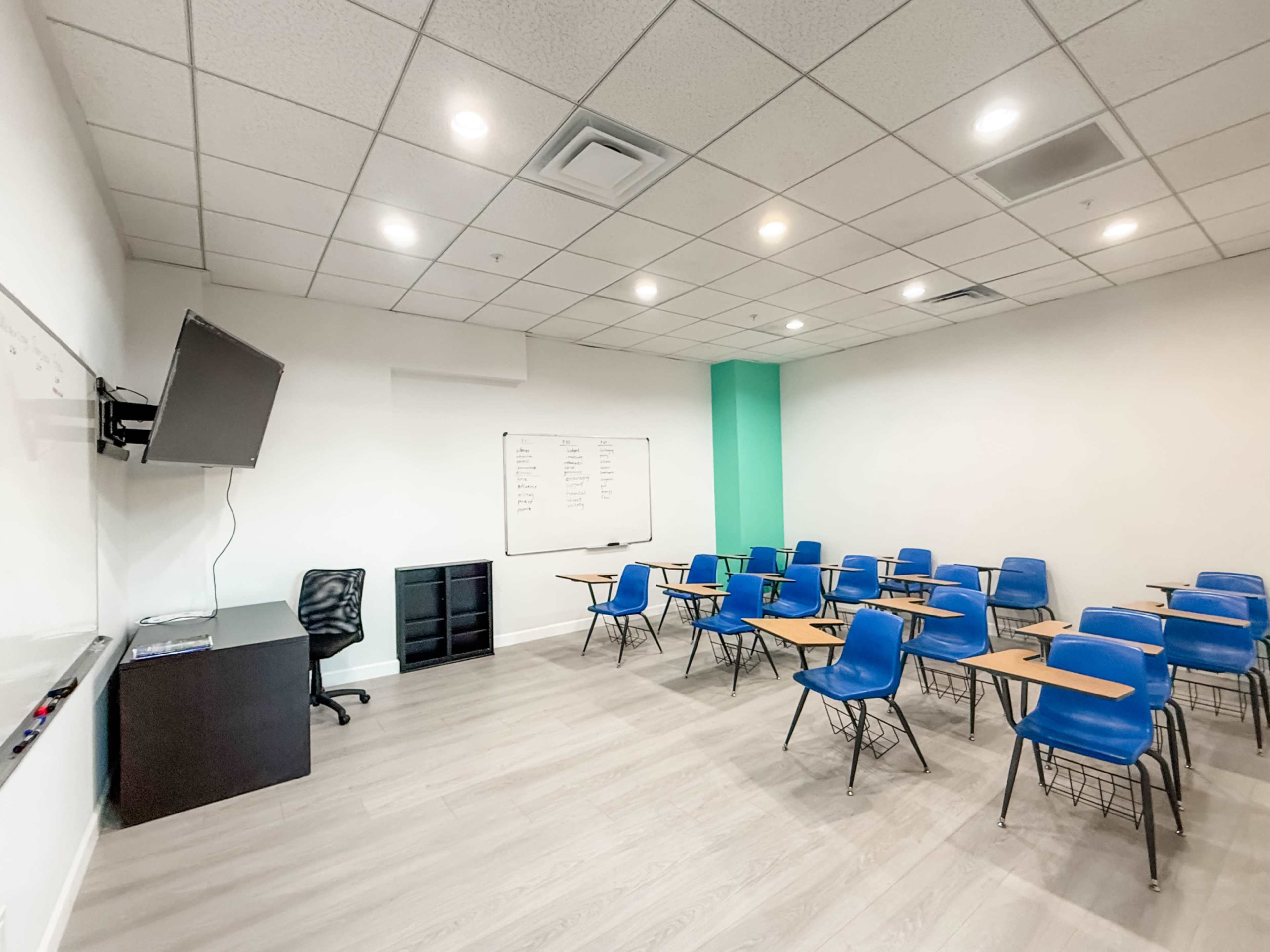 A classroom features blue student desks arranged in rows, a large screen mounted on the wall, and a whiteboard in front.