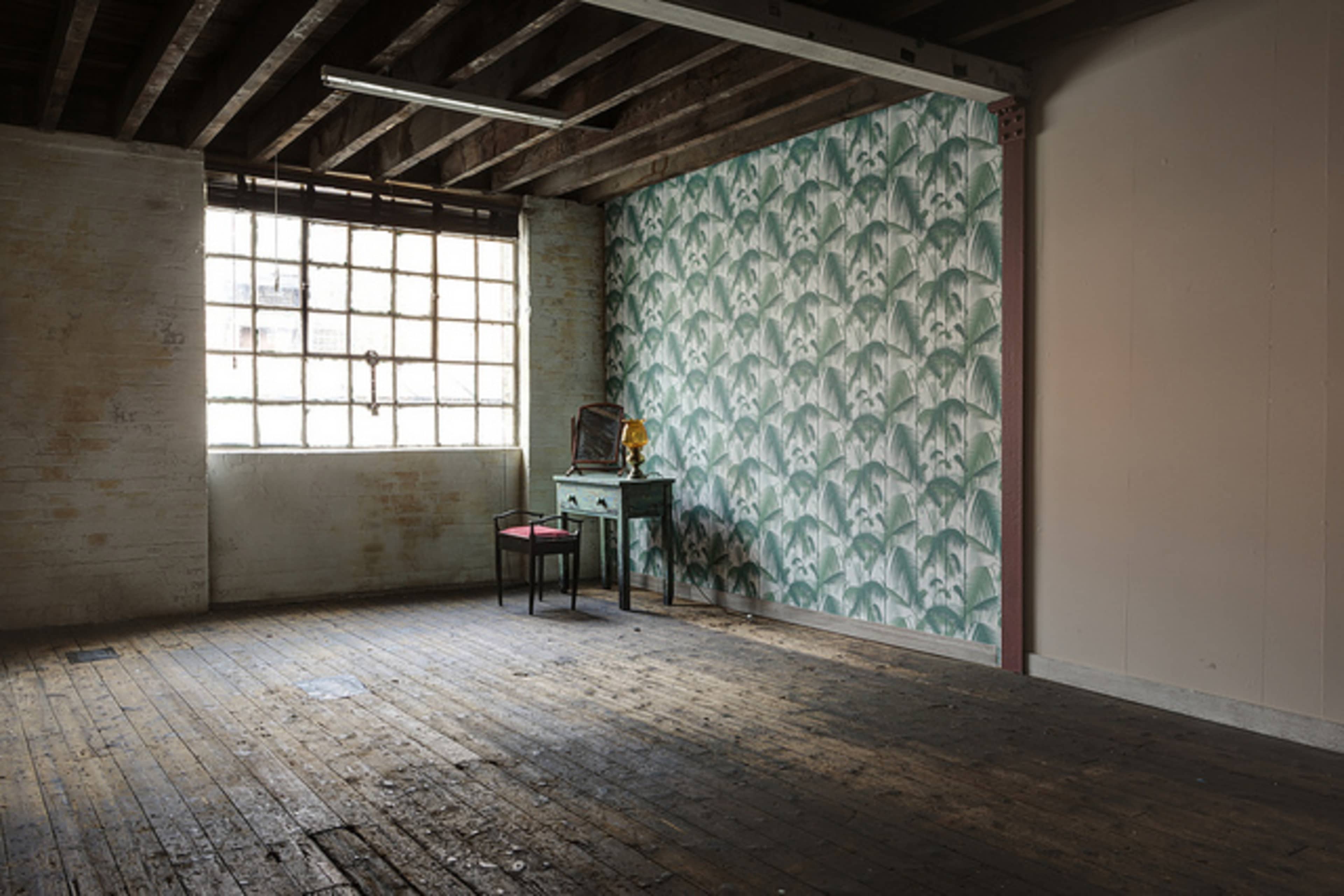 An empty room features a patterned wallpaper on one wall, a small table, a chair, and a large window allowing natural light to enter.
