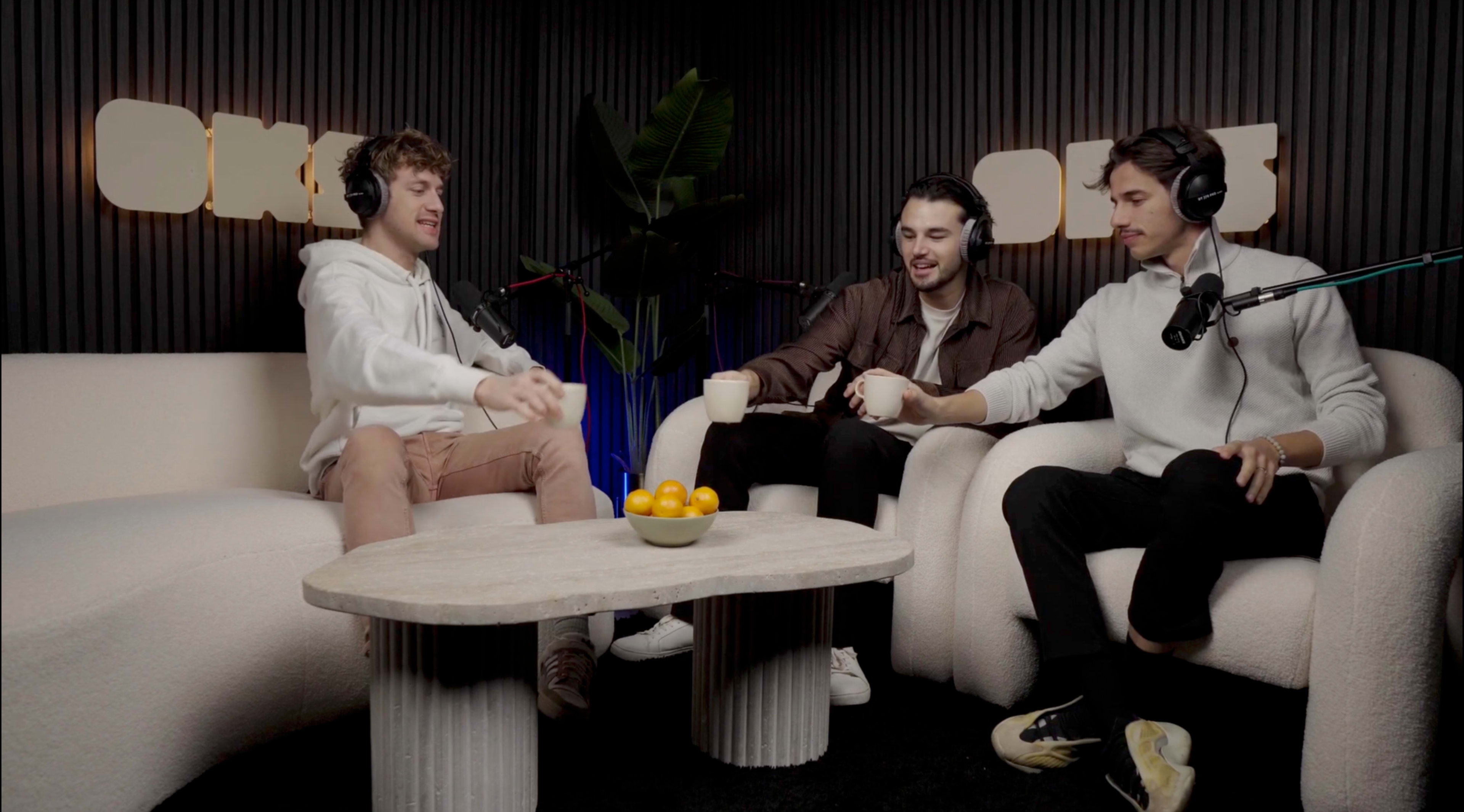 Three men sit on a plush white couch in a studio setting, engaging with each other while placing a bowl of oranges on a coffee table.